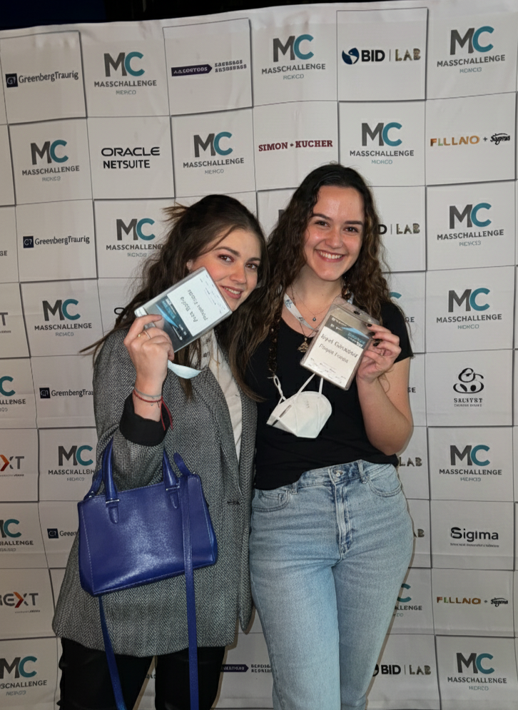 Dos mujeres jóvenes sonrientes en una conferencia, sosteniendo sus credenciales y una de ellas con un bolso azul, en un fondo con logotipos de patrocinadores y el evento MassChallenge.