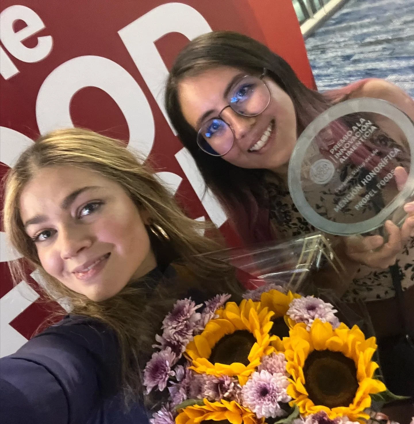 Dos mujeres sonriendo, una sostiene un trofeo y un ramo de girasoles y flores moradas, en un evento de premiación.