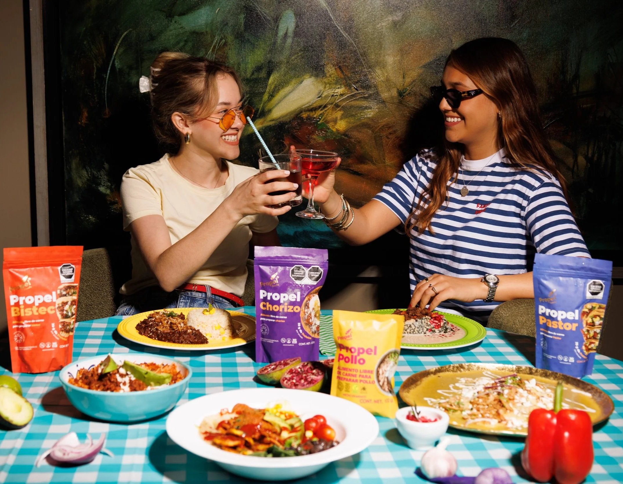 Dos mujeres disfrutando una comida y brindando con bebidas en un entorno festivo con varias comidas mexicanas y productos de carne en la mesa.