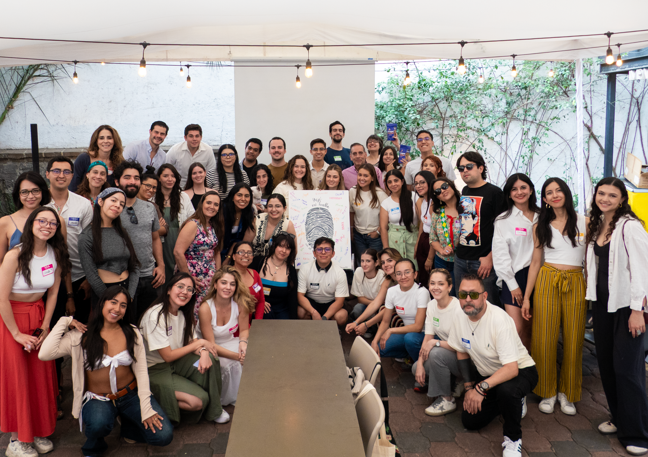 Un grupo grande de personas en una reunión al aire libre, algunas con gafas de sol, sosteniendo una cartel con un dibujo de una huella digital y escritos en español. El grupo parece feliz, con sonrisas y posando para la foto."