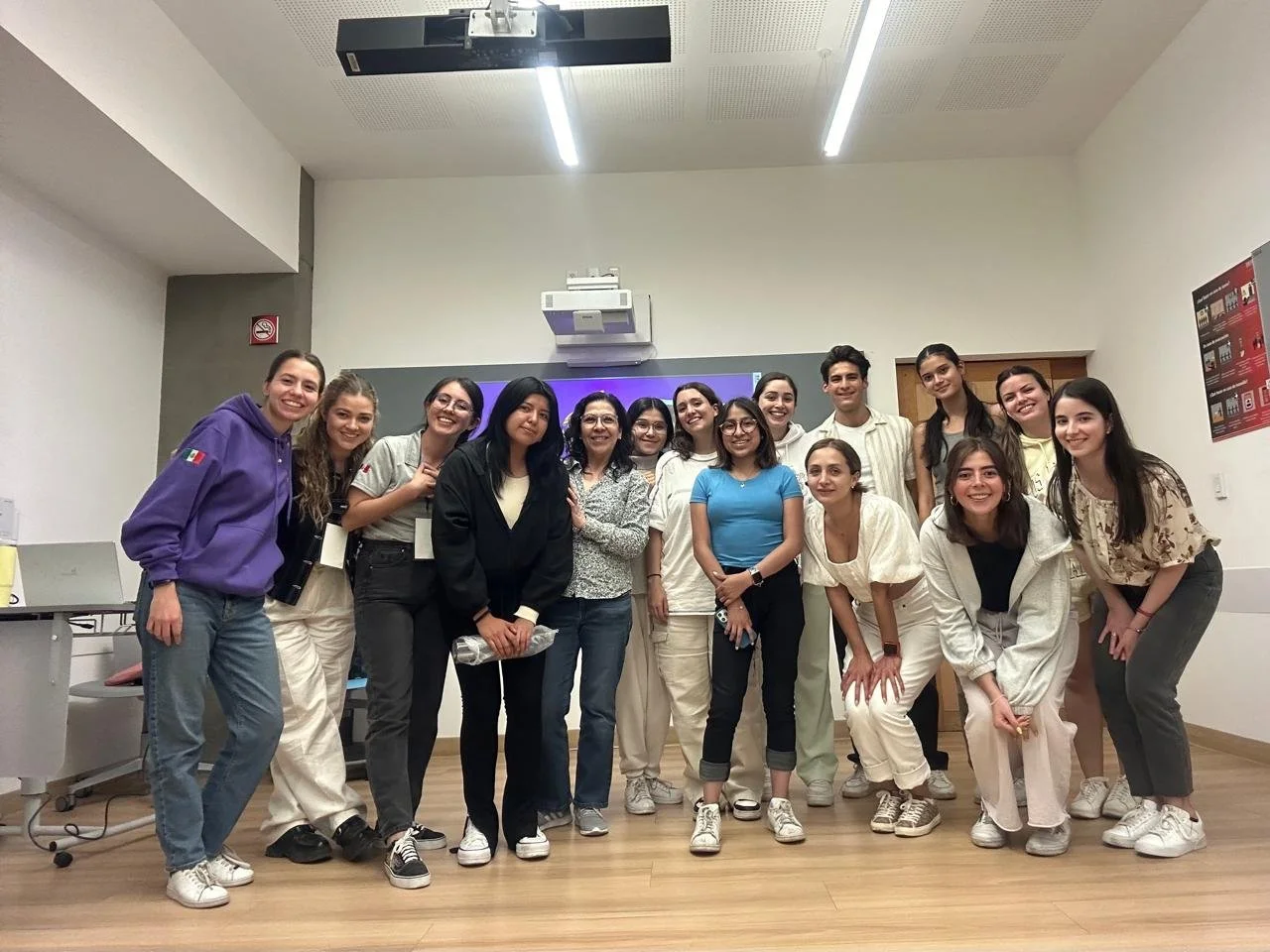 Grupo de personas posando en un aula, algunas con camisetas blancas, otras con ropa casual, frente a una pizarra y equipo audiovisual.