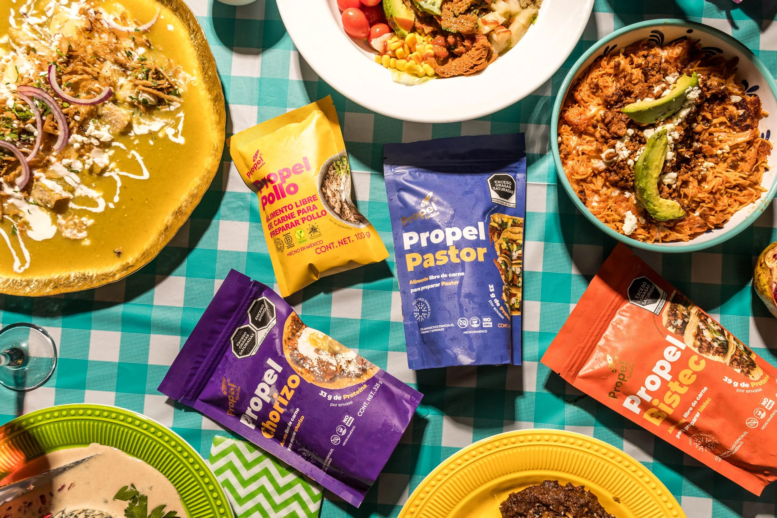 Imagen de comida mexicana en una mesa con diferentes salsas en paquetes Propel, platos con carne, pasta, frijoles y tortillas, sobre manteles a cuadros.