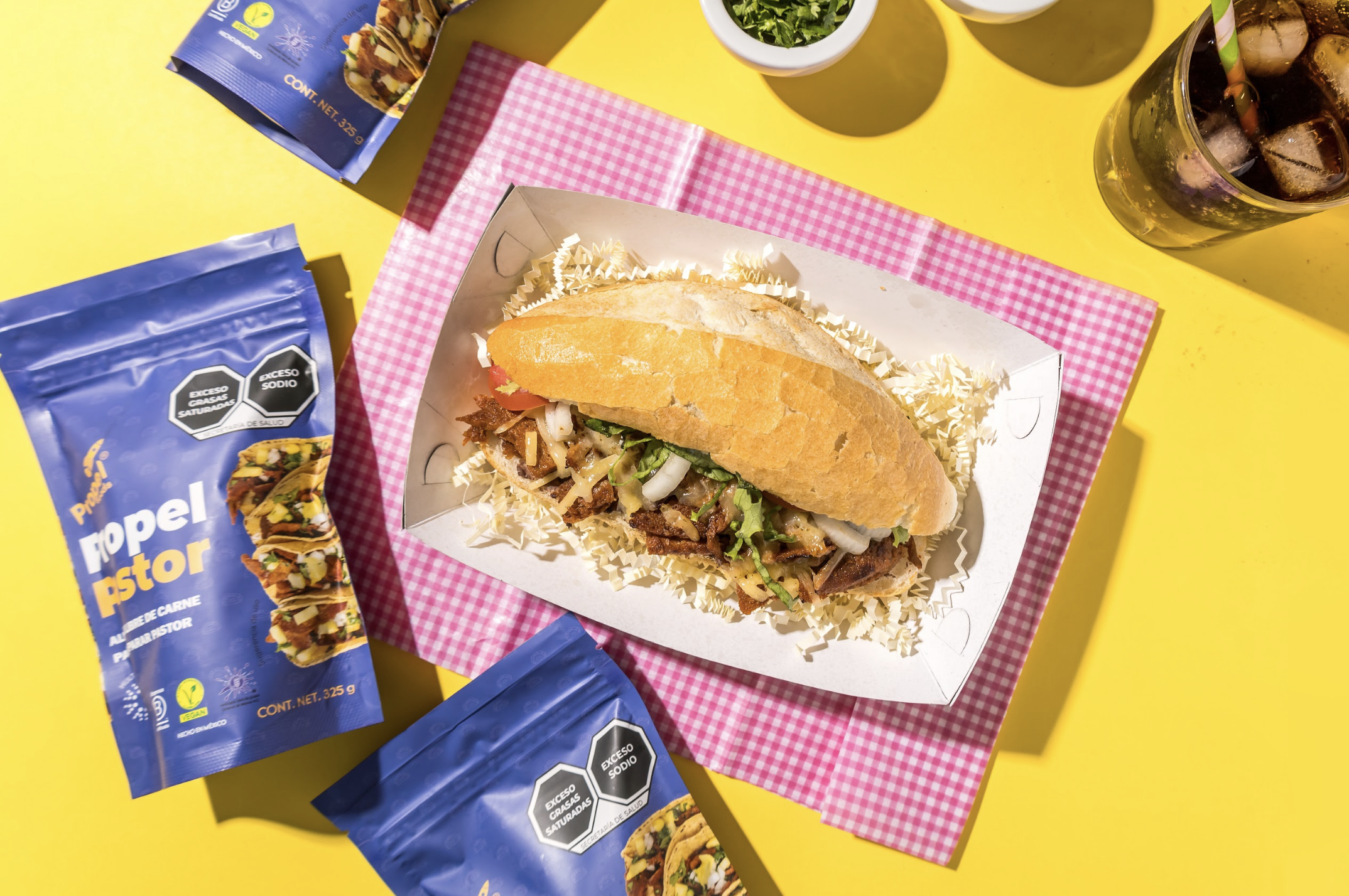 Bocadillo de carne con lechuga y cebolla en un pan, en una bandeja con papel decorativo, rodeado de paquetes de condimento en una mesa amarilla brillante.