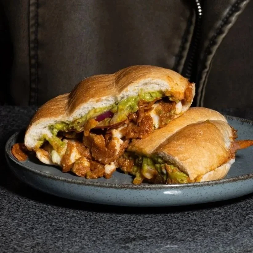 Torta de pastor plant based con guacamole, y otros ingredientes en un pan crujiente, sobre plato negro.