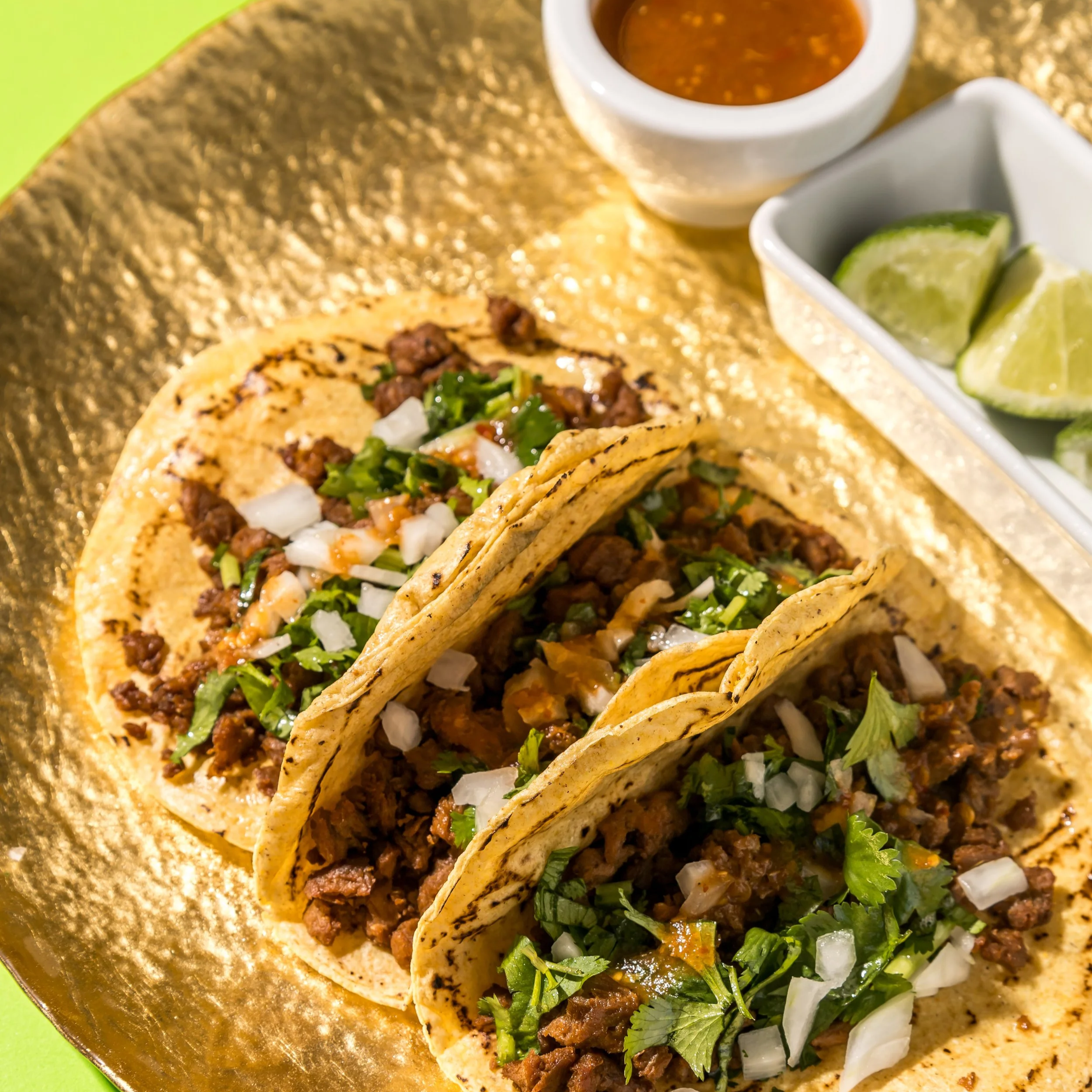 Tres tacos de bistec vegano con cebolla, cilantro y salsa, acompañados de limones y una salsa