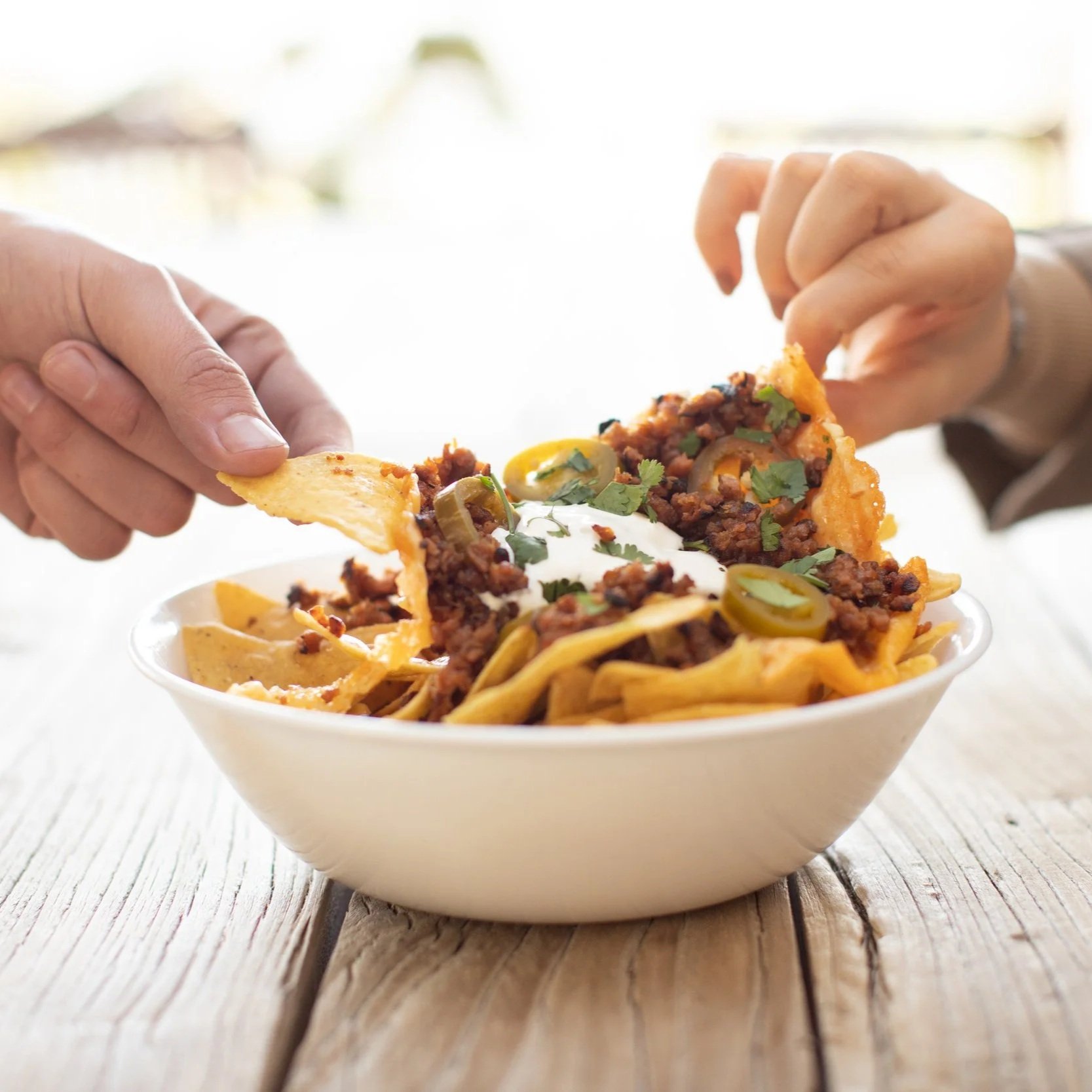 Persona comiendo nachos con carne, queso, crema, cilantro y jalapeños en un bol blanco sobre una mesa de madera.