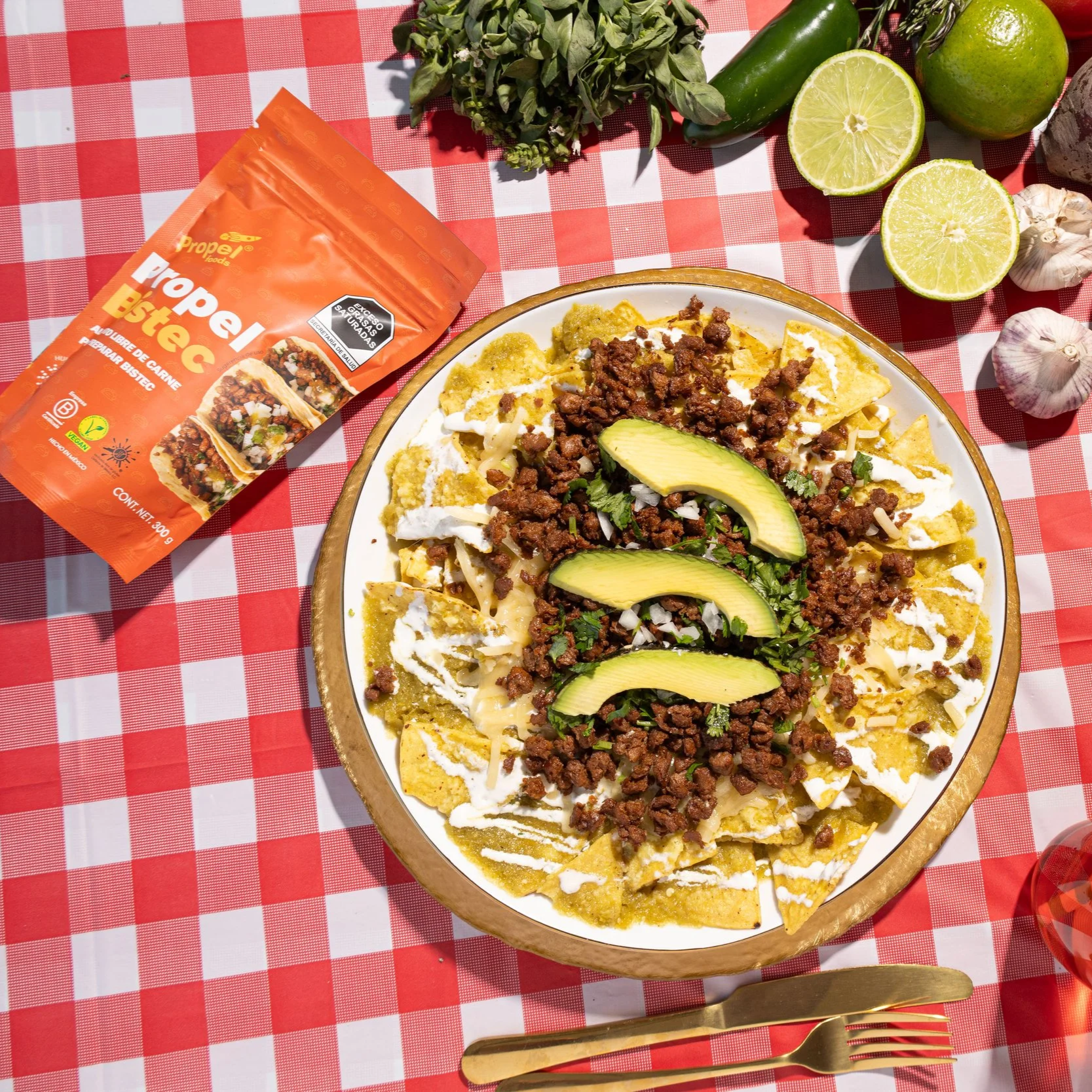 Plato de nachos cubierto con carne molida, rodajas de aguacate, crema, cilantro, y queso, en una mesa con una botella de especias, limones, ajo, y cubiertos dorados.