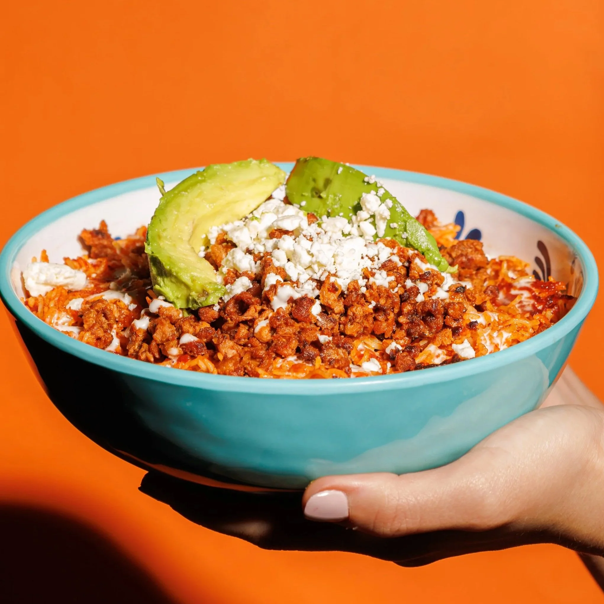 Un tazón azul con comida de fideo seco con carne molida vegana, queso, rodajas de aguacate y crema en un fondo naranja