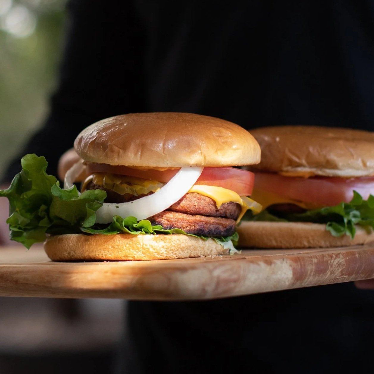 Dos hamburguesas veganas con pan tostado, carne, queso, lechuga, tomate, cebolla y pepinillos en una tabla de madera.