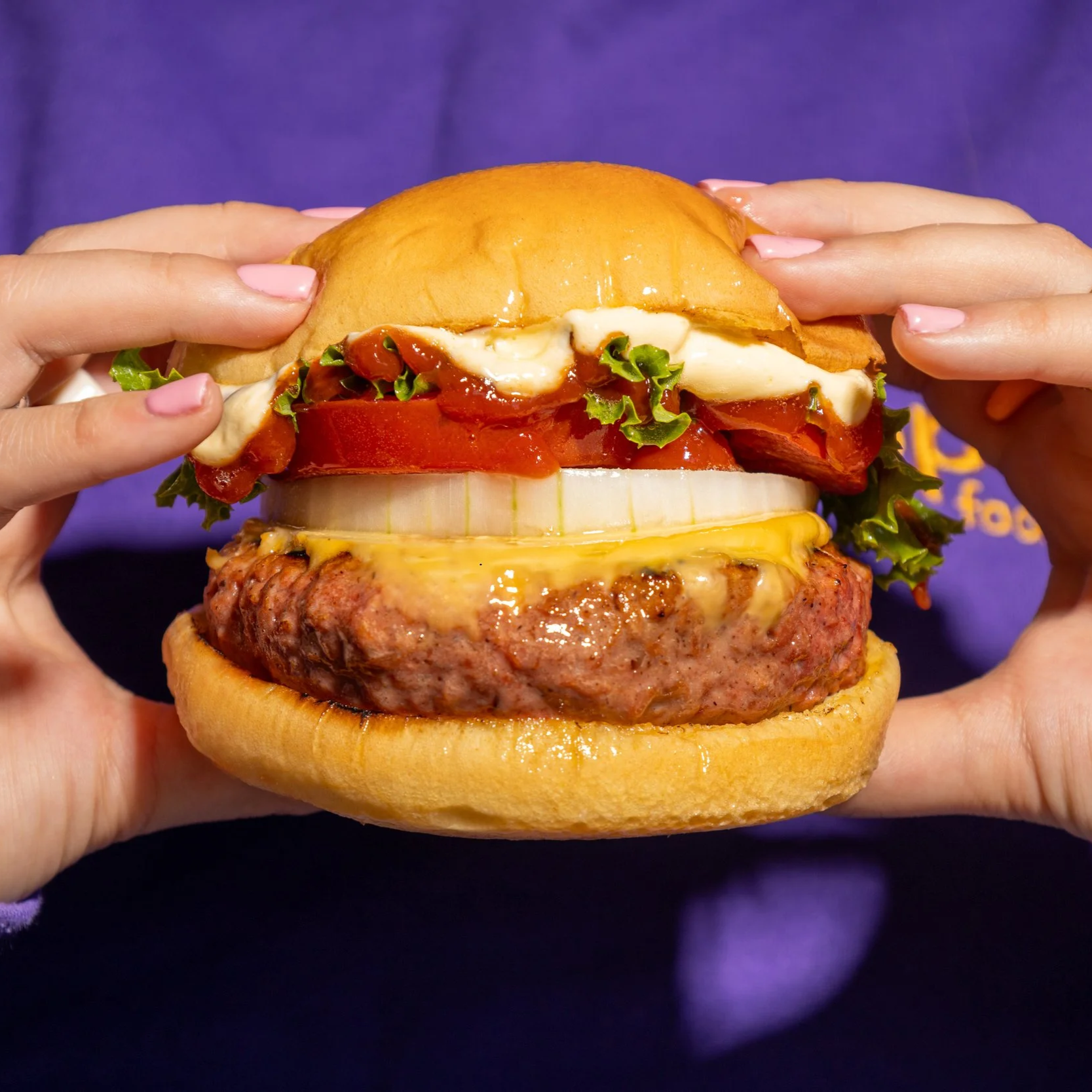 Primer plano de una hamburguesa con carne vegana, queso, cebolla, tomate, lechuga, salsa y pan, sostenida por dos manos con uñas pintadas de rosa.