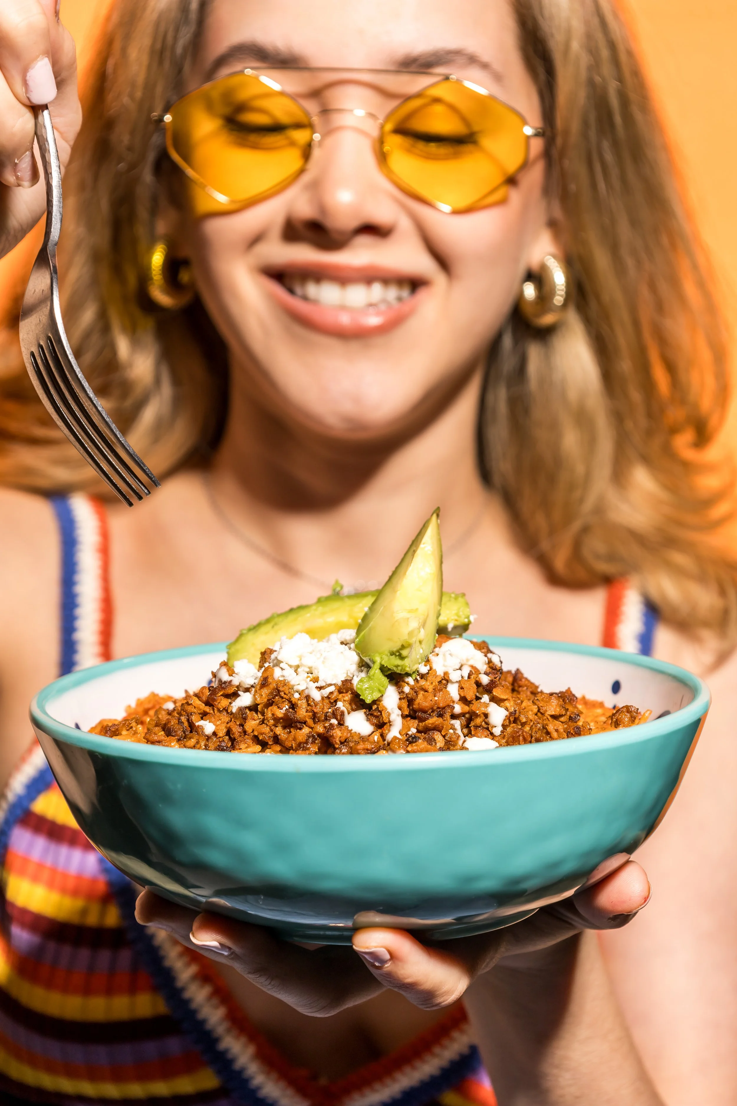 Mujer con gafas de sol amarillas, sosteniendo un bol de comida con aguacate y carne molida vegana, sonriendo.