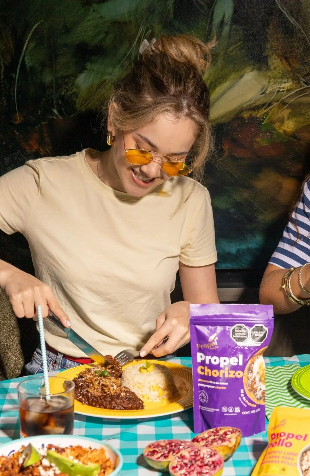 Una mujer con gafas amarillas y cabello recogido disfrutando de una comida mexicana, rodeada de alimentos y una bolsita de propel chorizo en una mesa con mantel a cuadros, en un ambiente festivo.