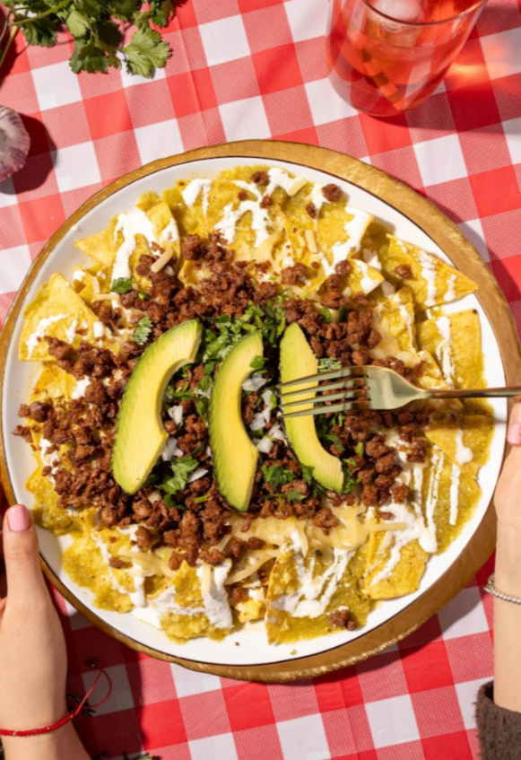 Plato de chilaquiles con bistec plant based, guacamole, cilantro, crema y salsa en un plato grande sobre una mesa con mantel de cuadros rojos y blancos.