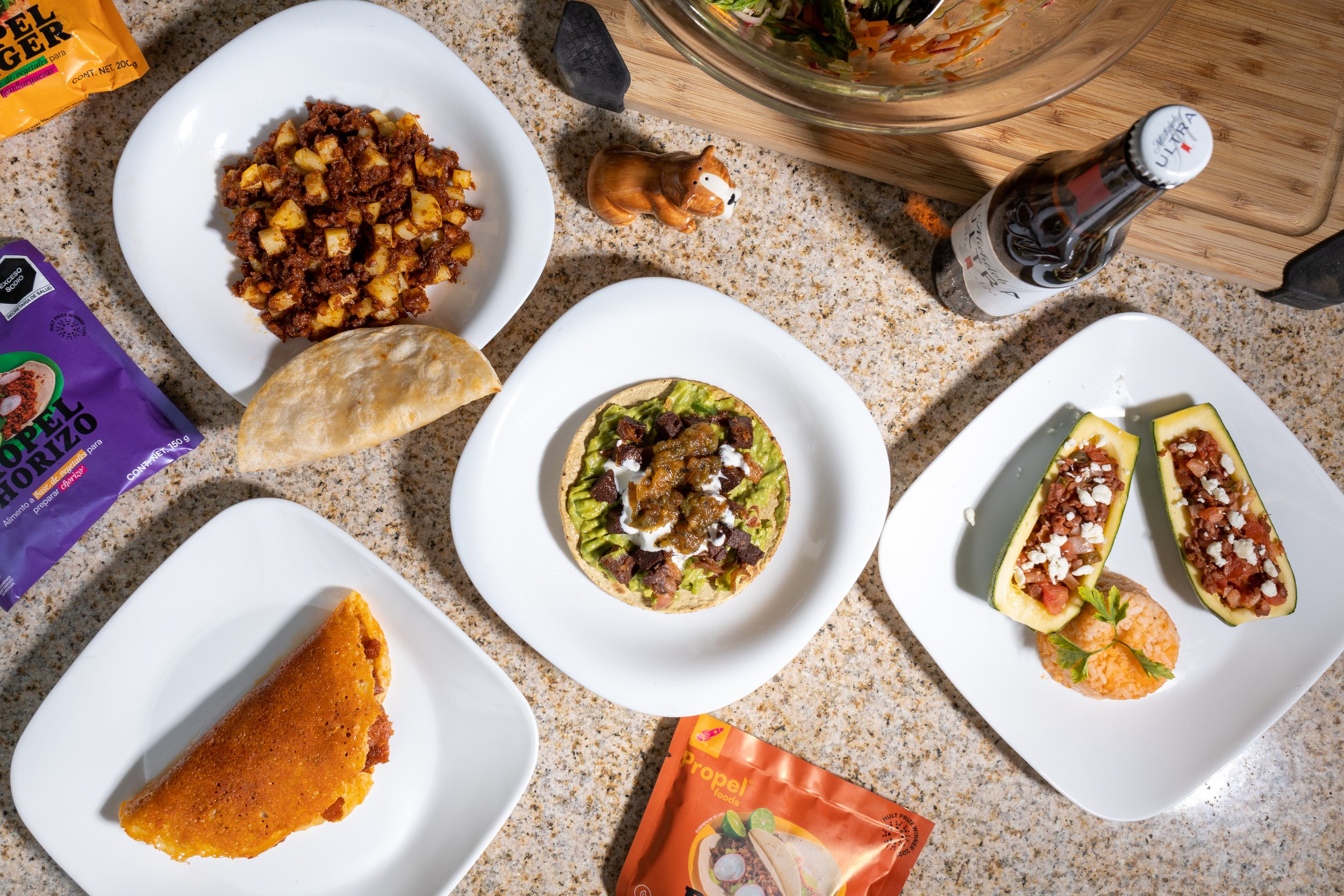 Variedad de platos mexicanos: un taco con guacamole, dos calabacitas rellenas, una quesadilla, y una enchilada, acompañados de una sopa, una bebida y snacks.