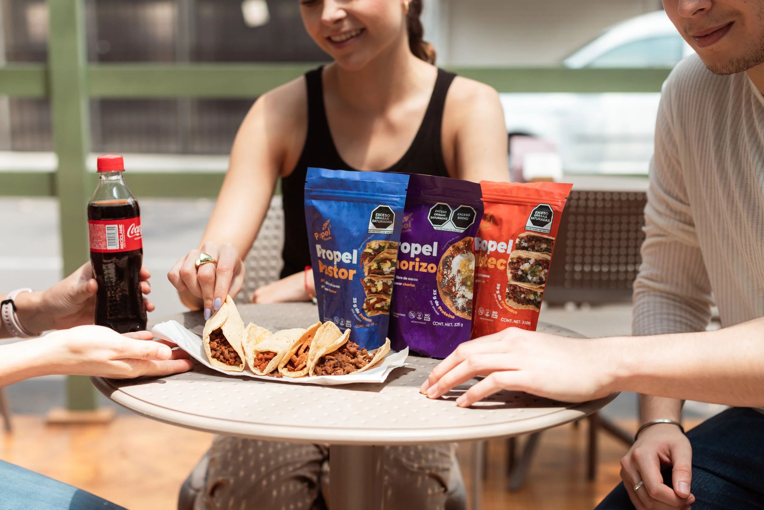 Tres personas sentadas alrededor de una mesa compartiendo tacos de carne. En el centro hay bolsas de carne para tacos de la marca Propel, y un vaso de refresco Coca-Cola.