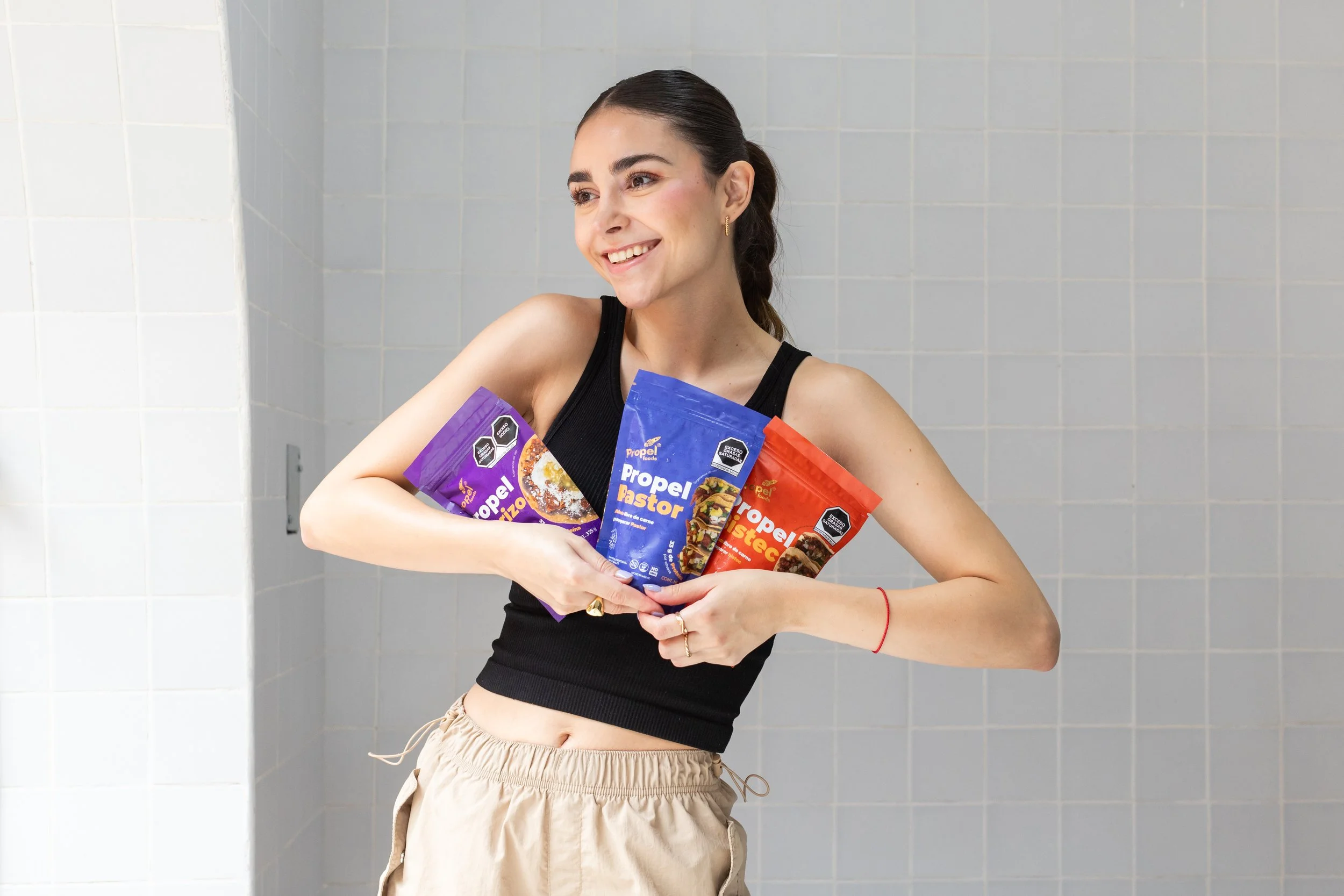 Joven mujer sonriendo sosteniendo paquetes de producto alimenticio en un fondo de azulejos blancos.