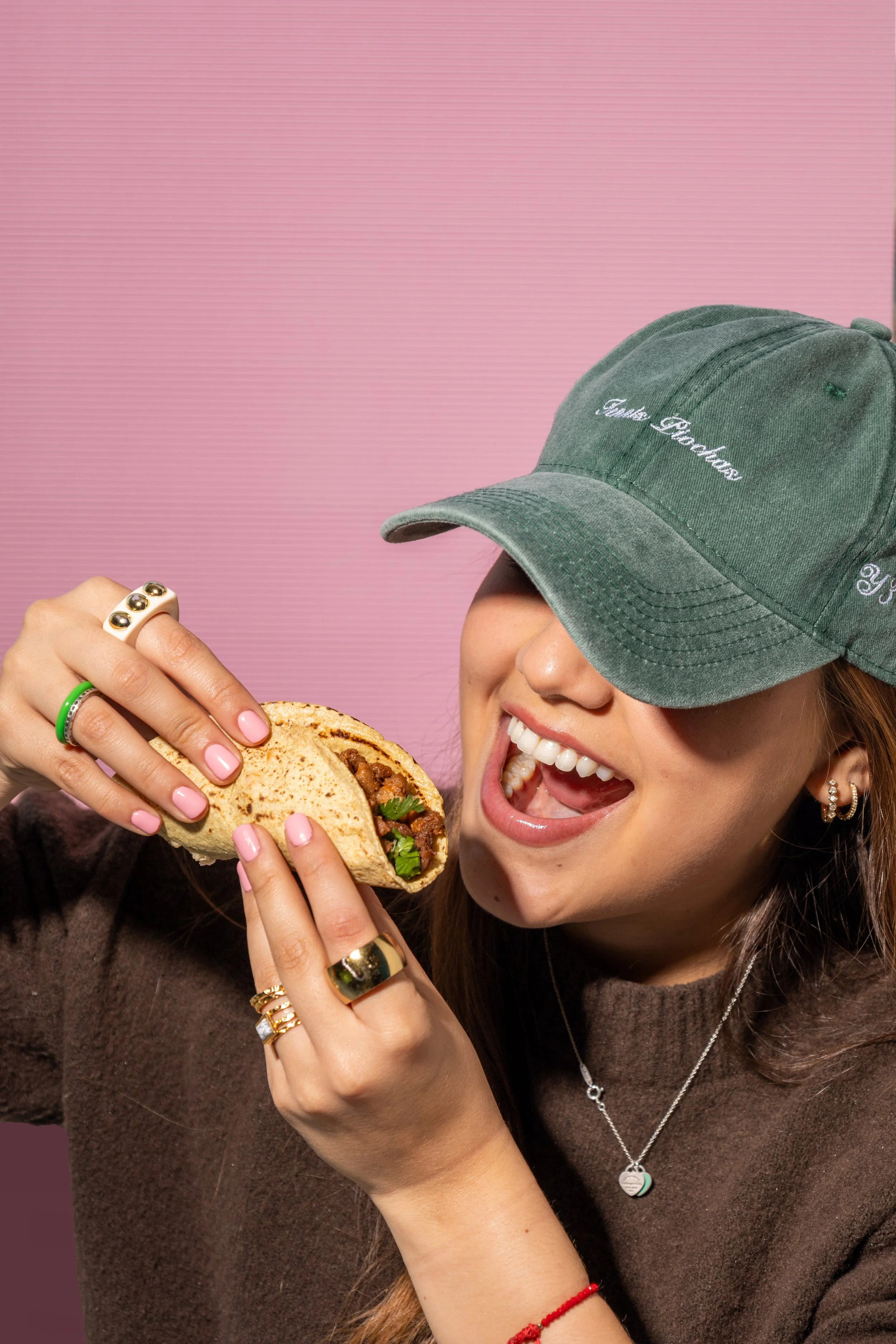 Una mujer sonriente sosteniendo un taco en una mano, usando una gorra verde y ropa marrón, con accesorios de joyería y fondo rosa.