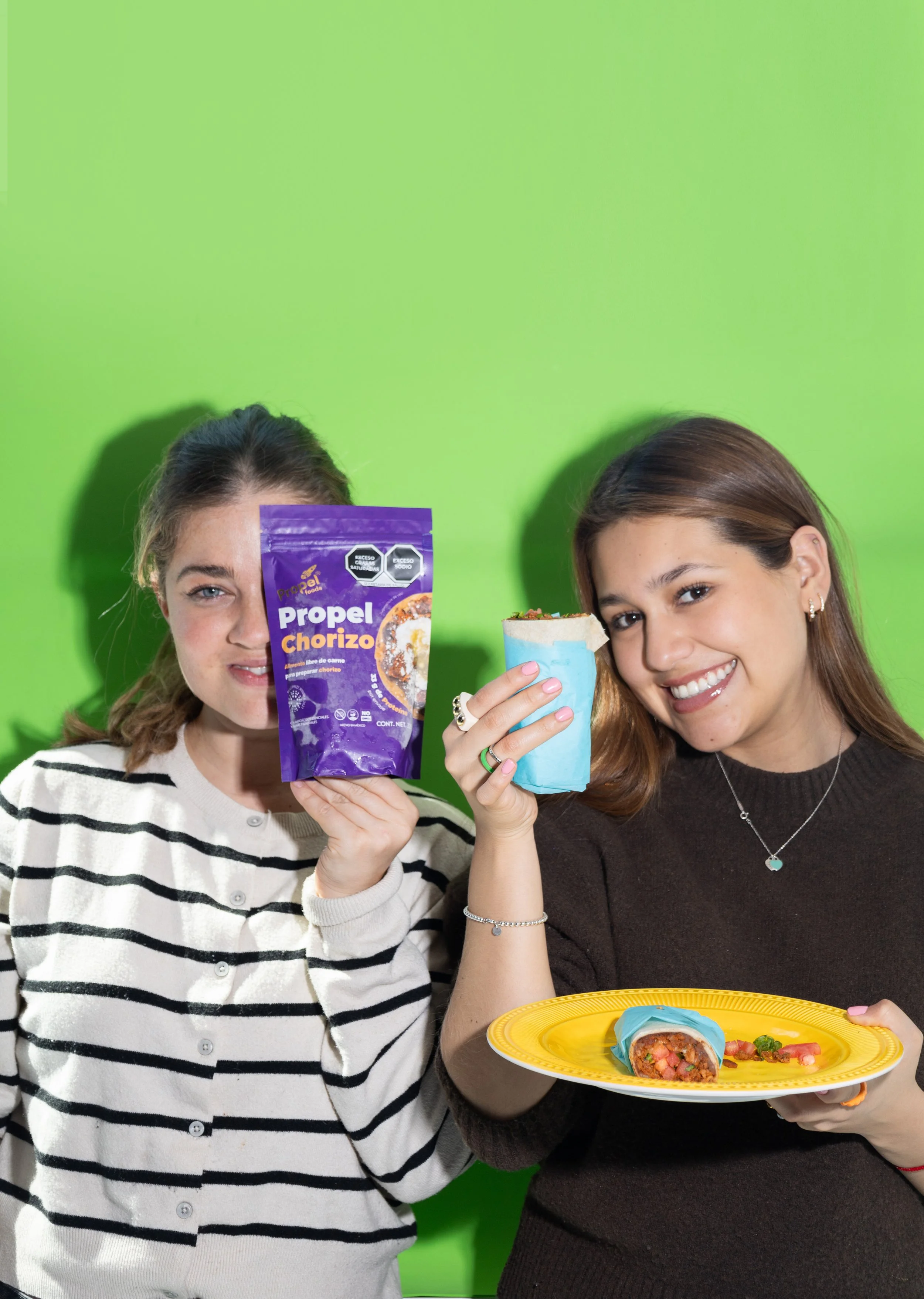 Dos jóvenes sosteniendo comida mexicana, una con un paquete de chorizo y la otra con un burrito, con fondo verde.