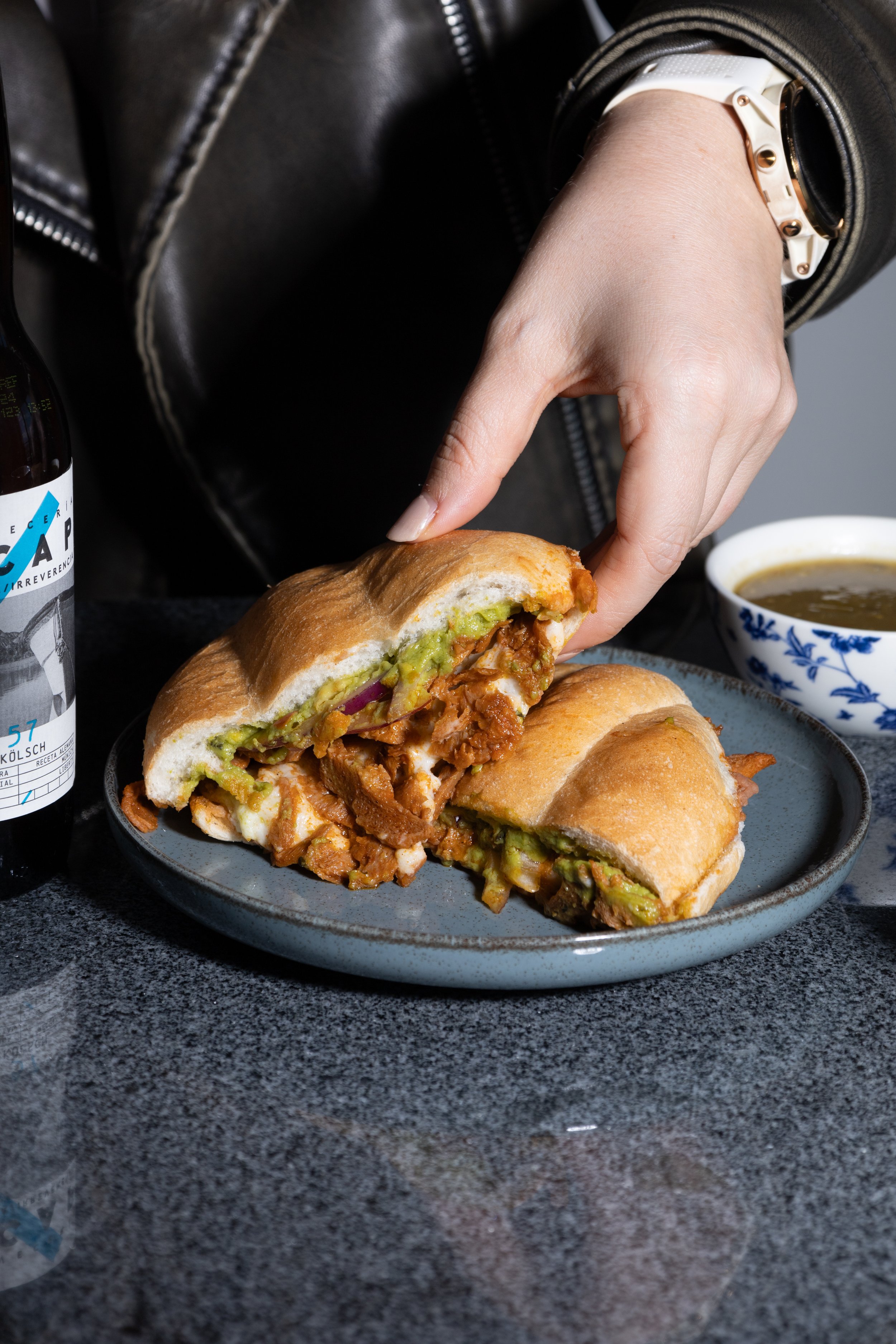 Una persona sosteniendo un sándwich de pan crujiente con carne, aguacate, y otros ingredientes, servido en un plato sobre una mesa. También hay una taza con sopa, una botella y un proceso de comer.