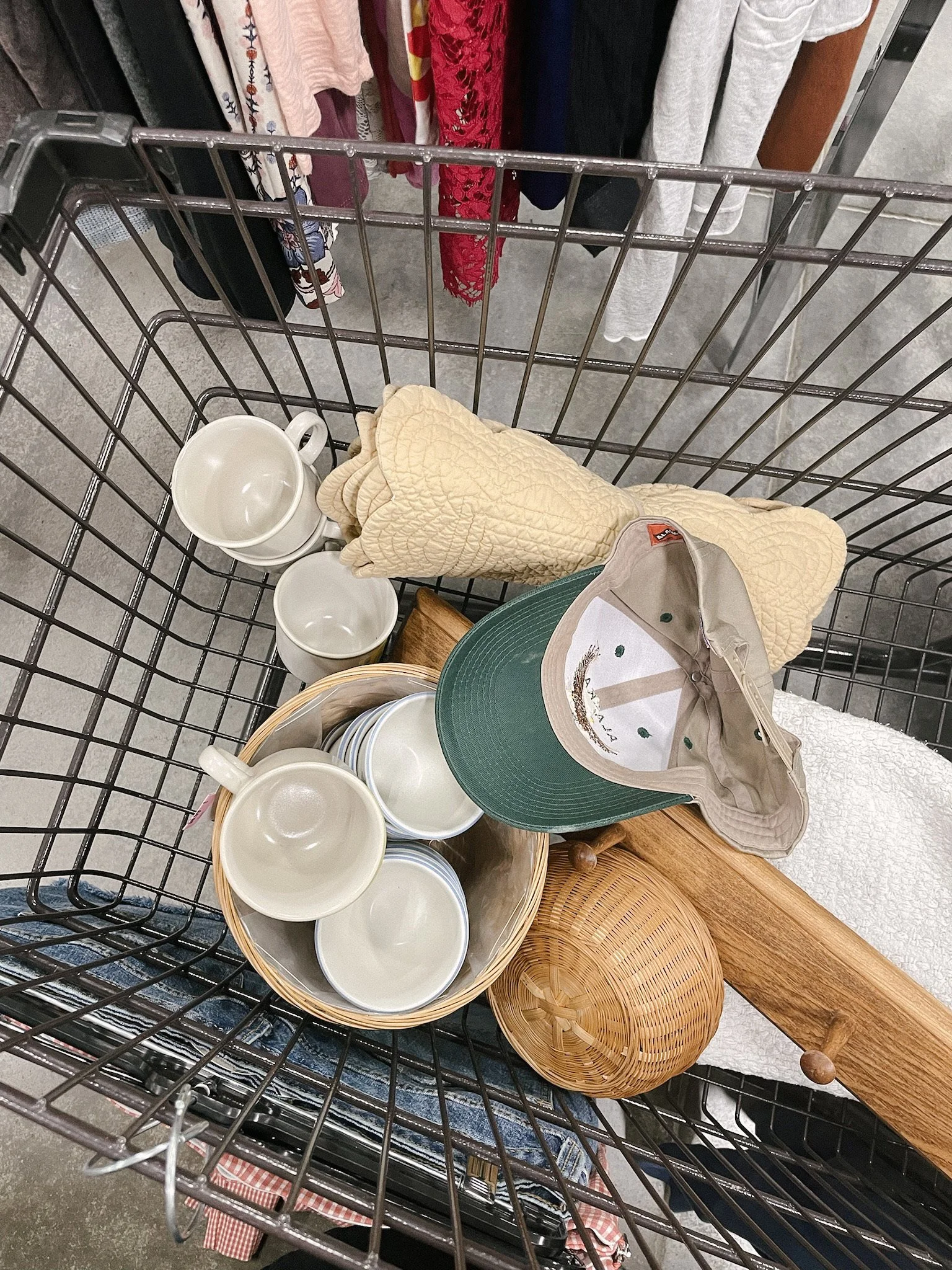 A shopping cart filled with ceramic mugs, a beige cap, a yellow quilted oven mitt, a round wooden container, and a green cloth hat, at a thrift store.