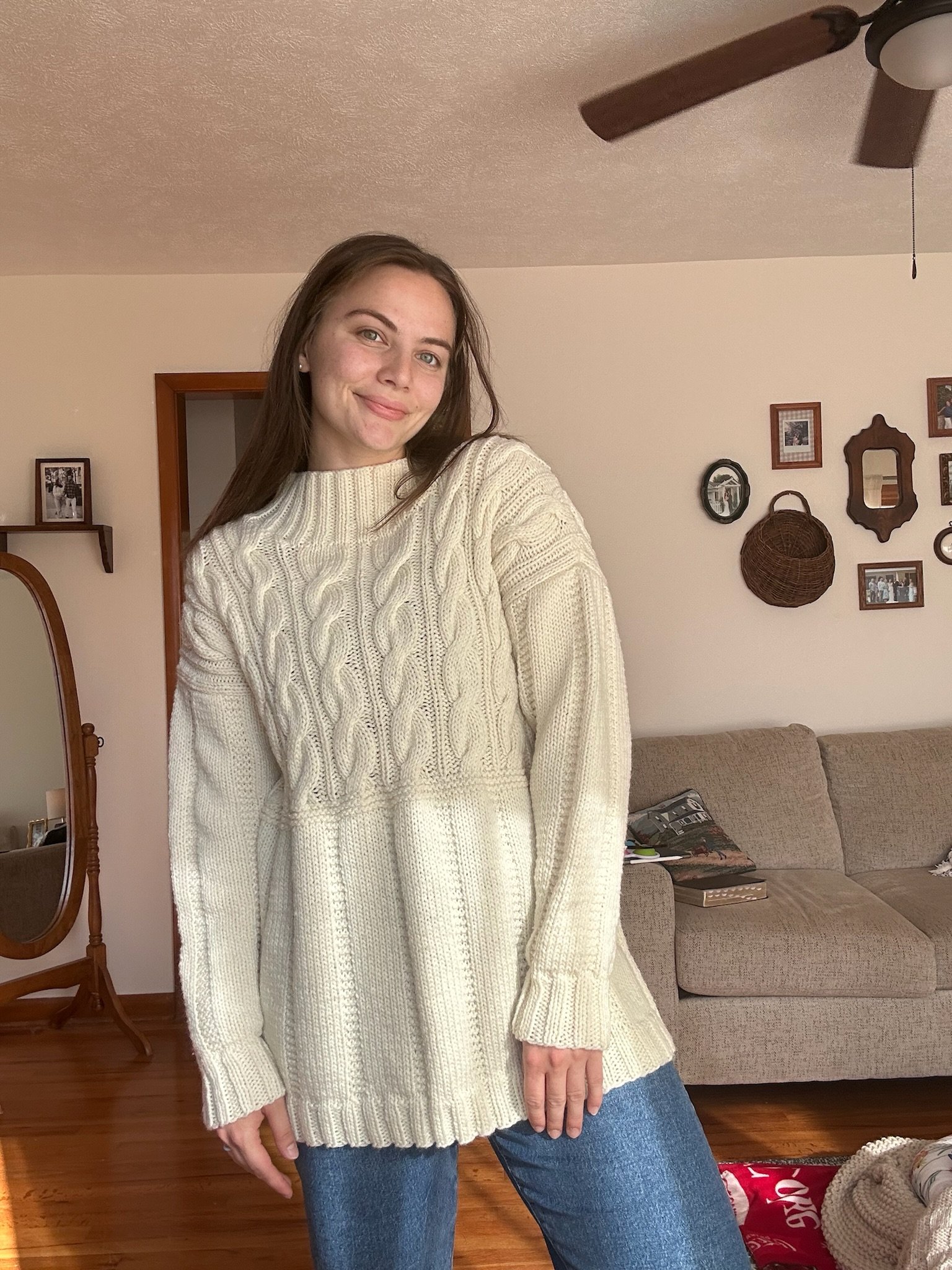 A young woman with long brown hair is standing in a living room, wearing a cream-colored knitted sweater and blue jeans, smiling at the camera.