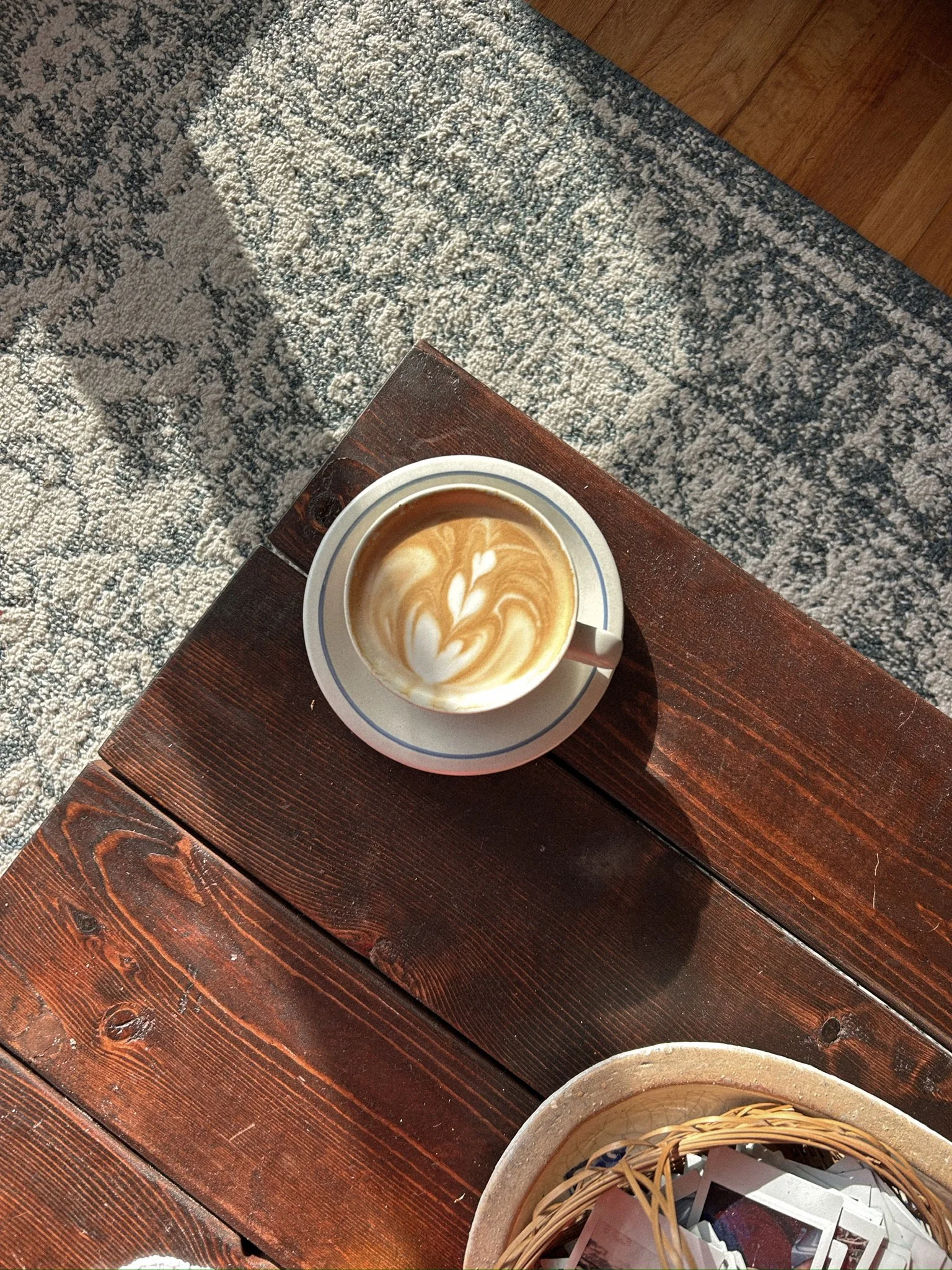 A cup of coffee with latte art on a wooden table, with sunlight casting a shadow. Part of a basket with papers is visible in the lower part of the image.