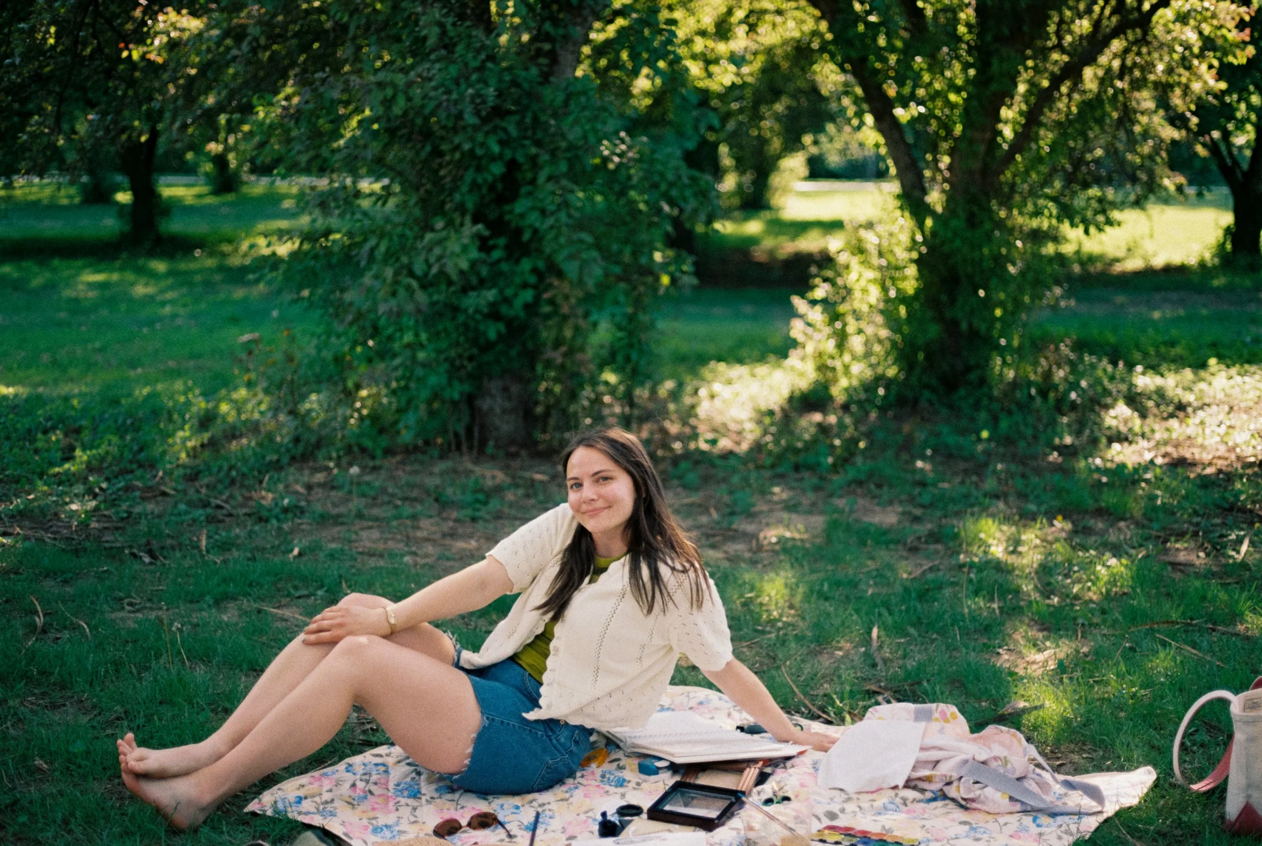 A young woman relaxing on a blanket in a park, surrounded by trees, with makeup and beauty products around her.