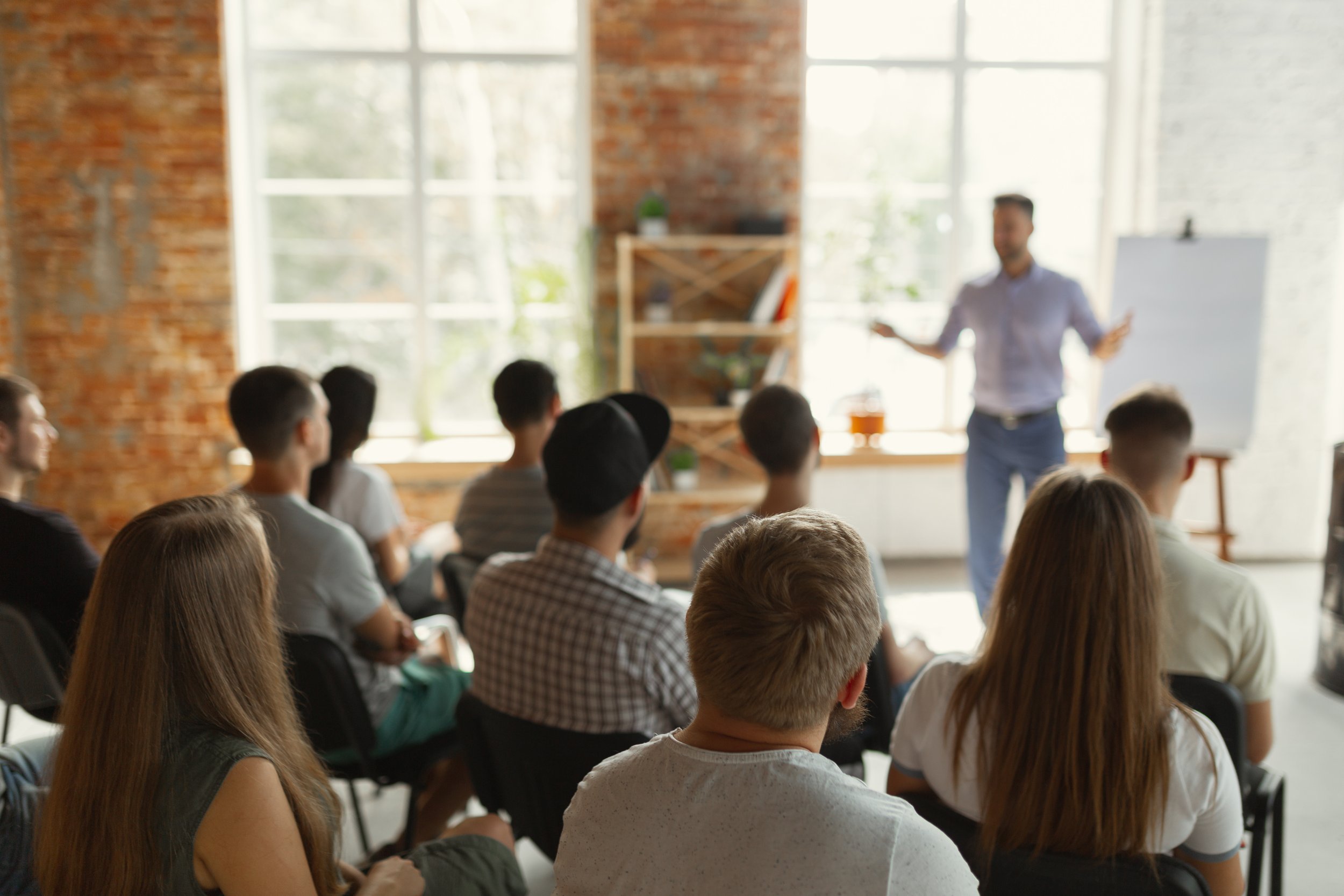 A man giving a presentation or lecture to a group of young adults seated in a bright, modern room with large windows and exposed brick walls.