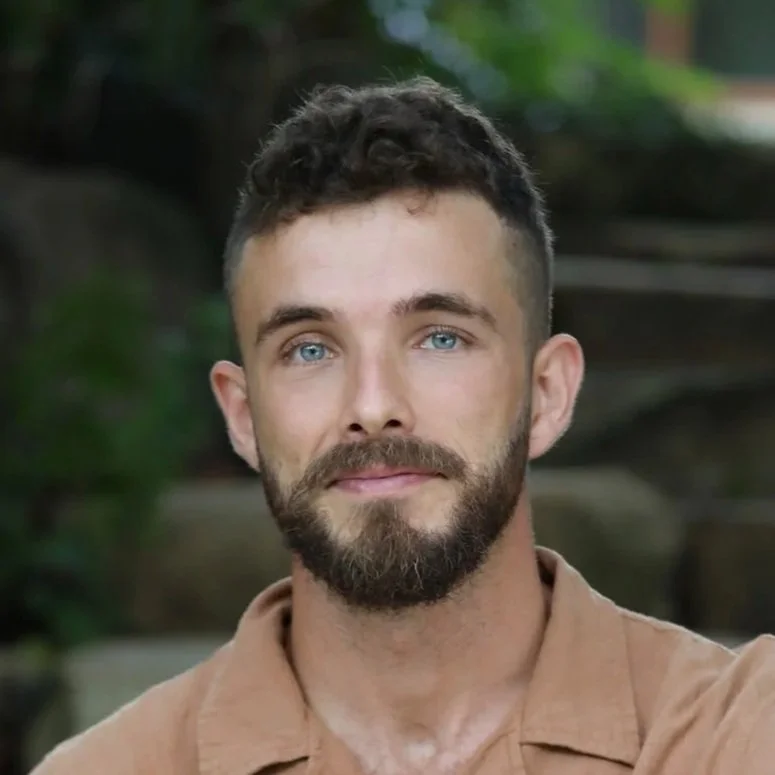 Close-up of a young man with blue eyes, dark curly hair, a beard, wearing a beige shirt, outdoors with greenery in the background.