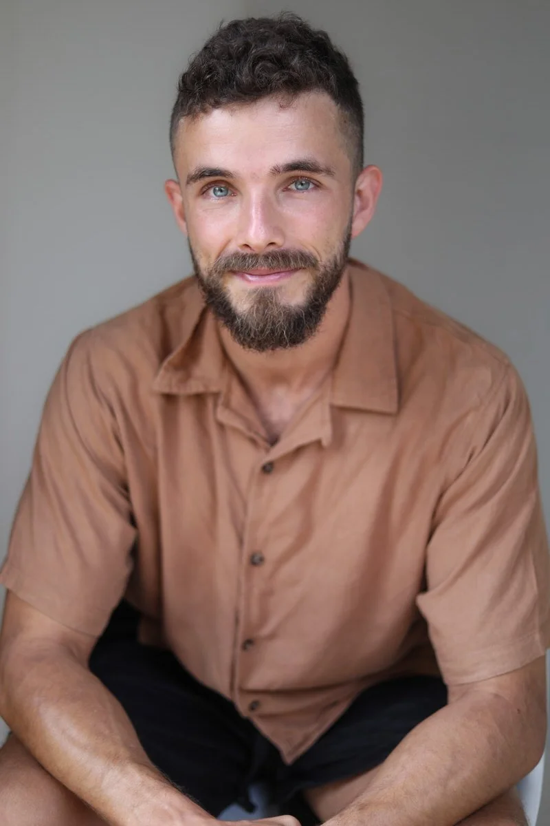A man with blue eyes, a beard, and short curly brown hair smiling while sitting against a gray background, wearing a brown short-sleeve button-up shirt.