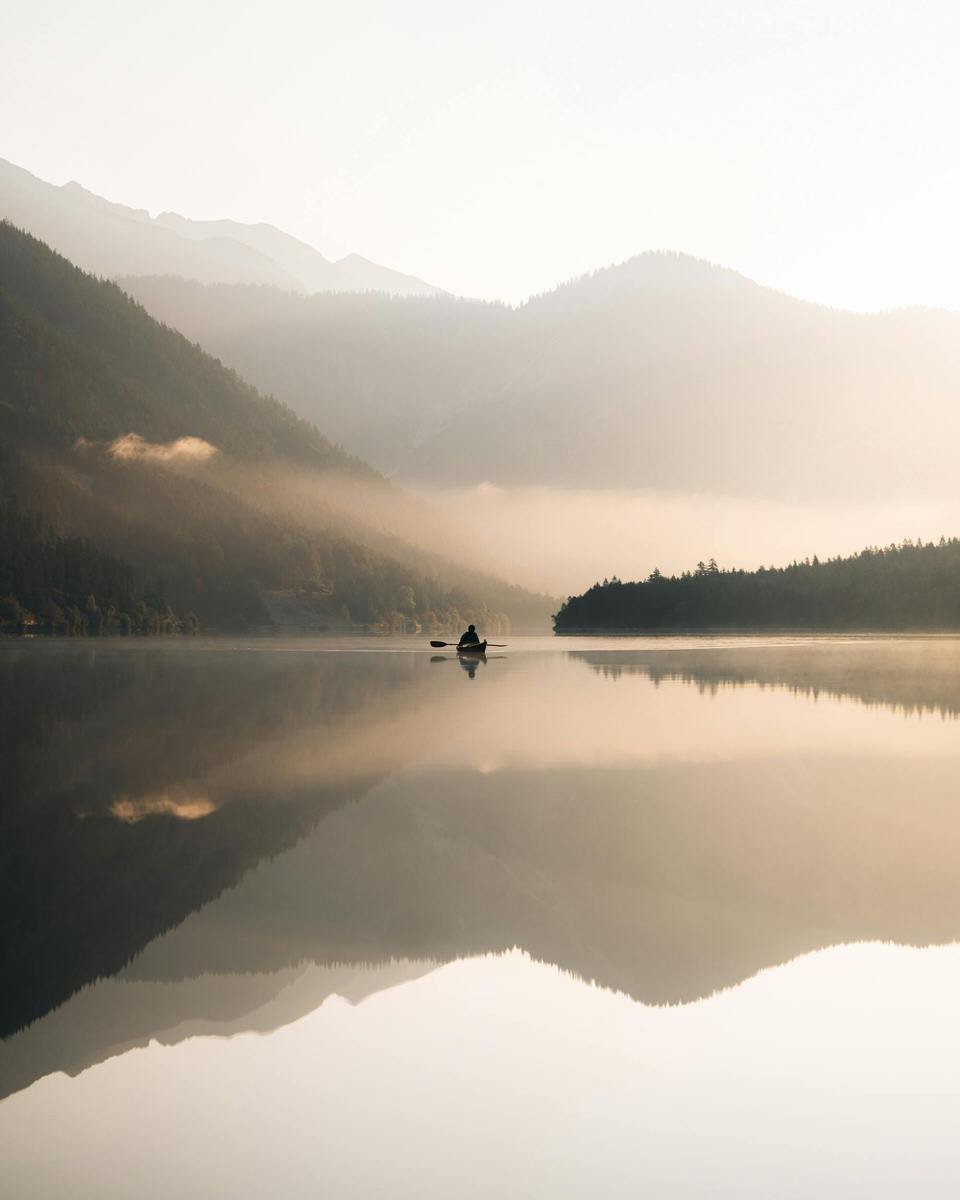 A person kayaking on a calm lake surrounded by misty mountains with reflections on the water.
