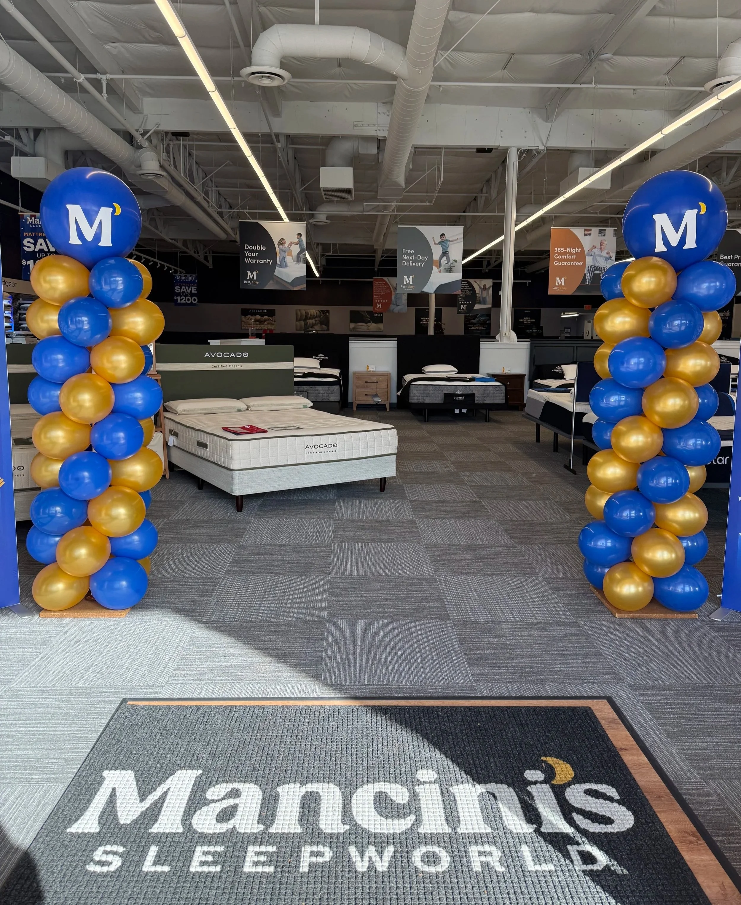Entrance to a mattress store with blue and gold balloons, showroom displaying various beds, and a mat with the Mancinis Sleepworld logo