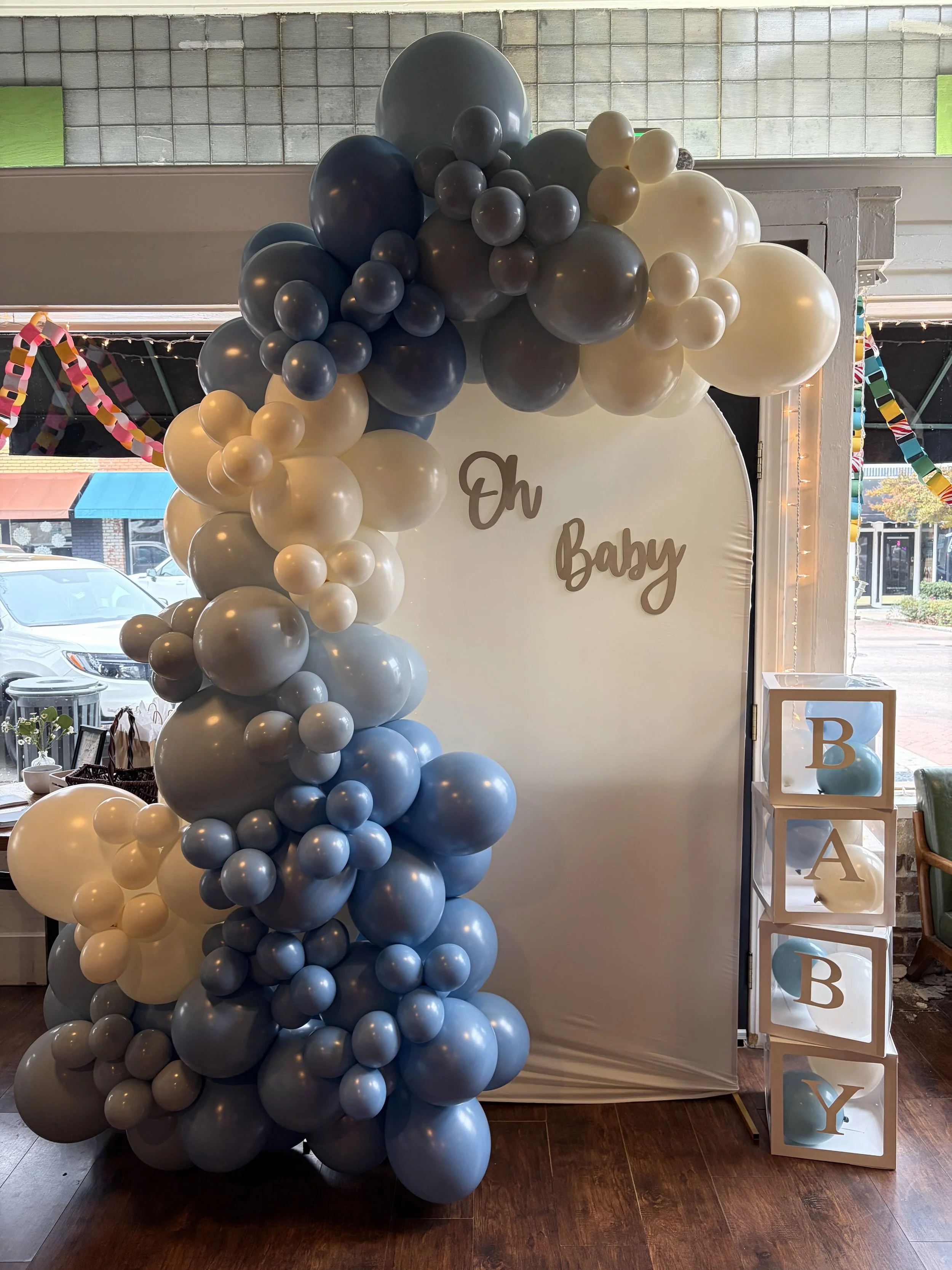 Decorative baby shower display with a large cluster of blue, gray, white, and cream balloons arranged around a white backdrop that reads 'Oh Baby.' The display also features cubes stacked vertically spelling out 'BABY' with blue balloons inside some 