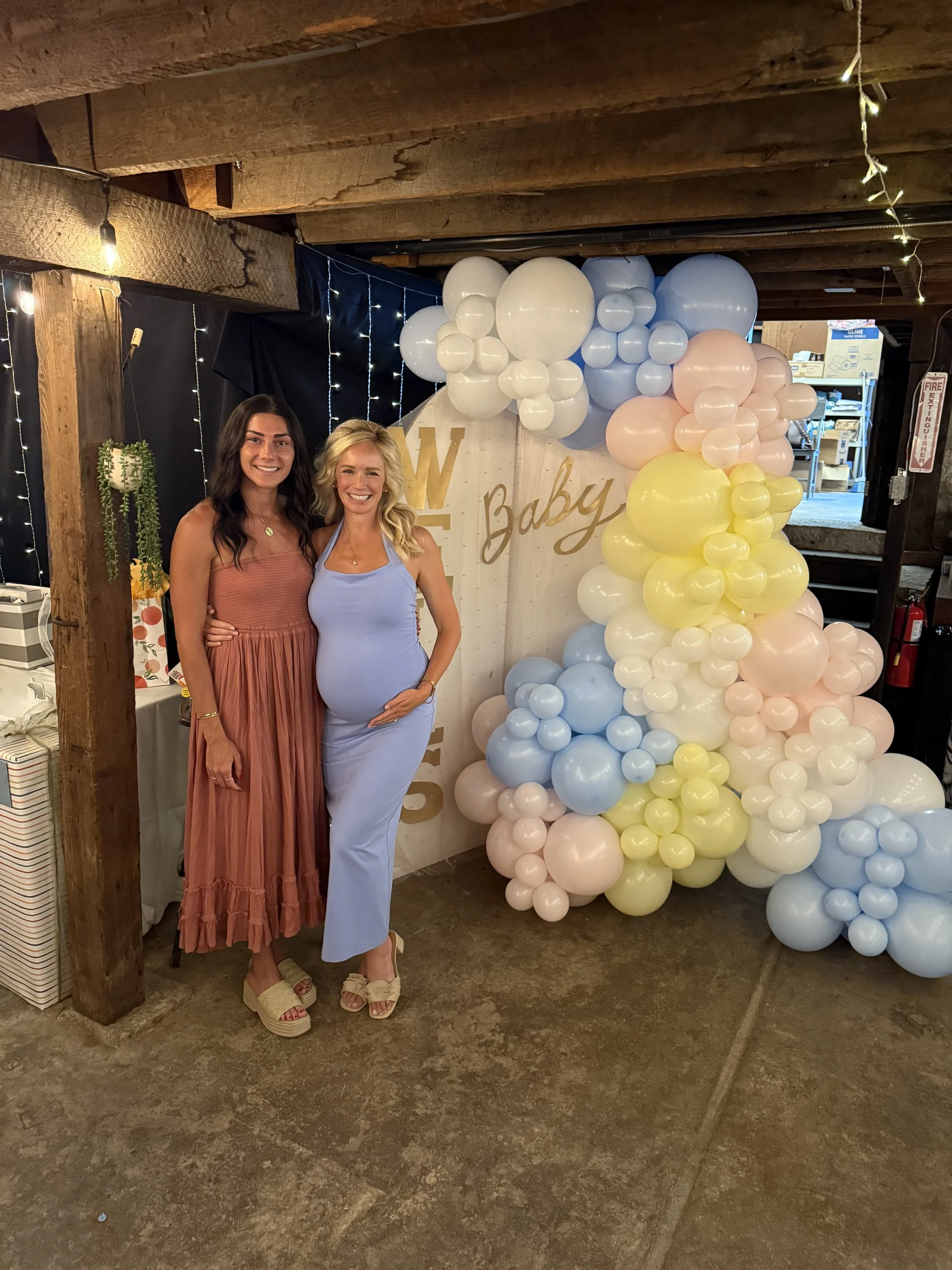 Two women standing together inside a decorated area with balloons and a sign that says "Baby". One woman is pregnant, with her hand on her belly, wearing a light blue dress. The other woman is wearing a rust-colored dress. There are string lights and
