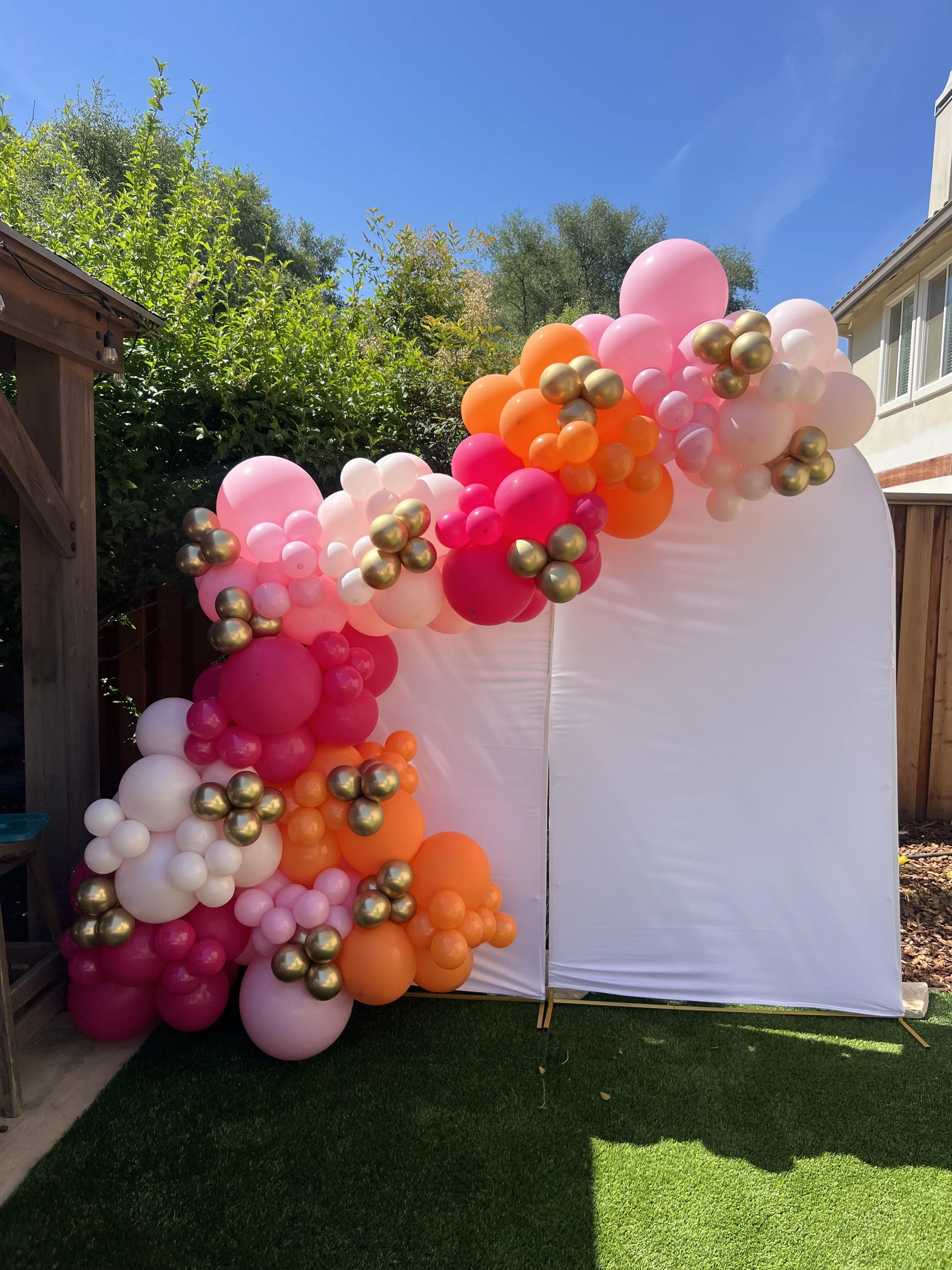 Colorful balloon arrangement with pink, orange, white, and gold balloons, set against a white backdrop outdoors with greenery and blue sky.