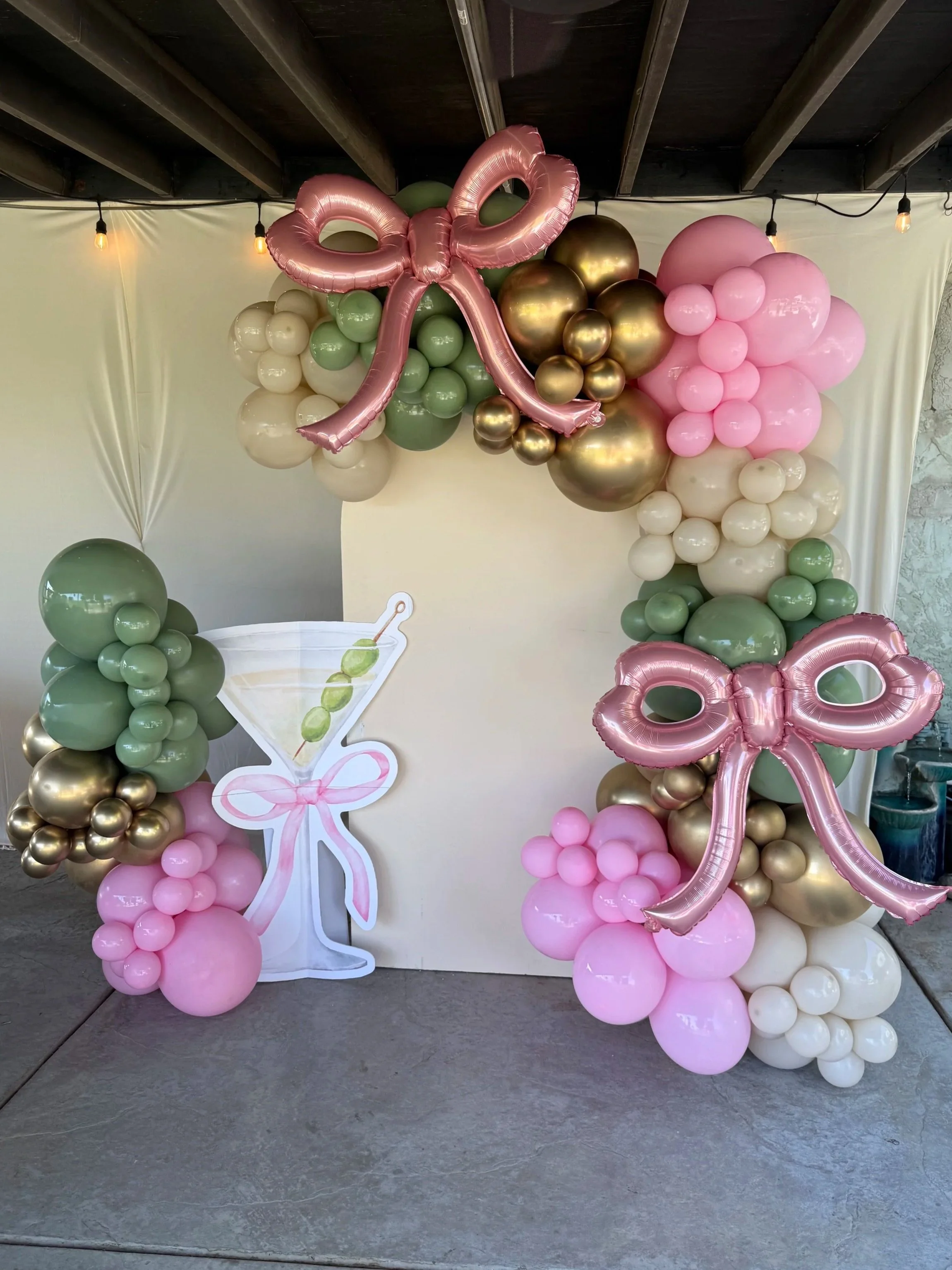 Decorative balloon arch with pink bows, gold, white, and green balloons, and a large cutout of a martini glass with olives and a pink ribbon in a party setting.