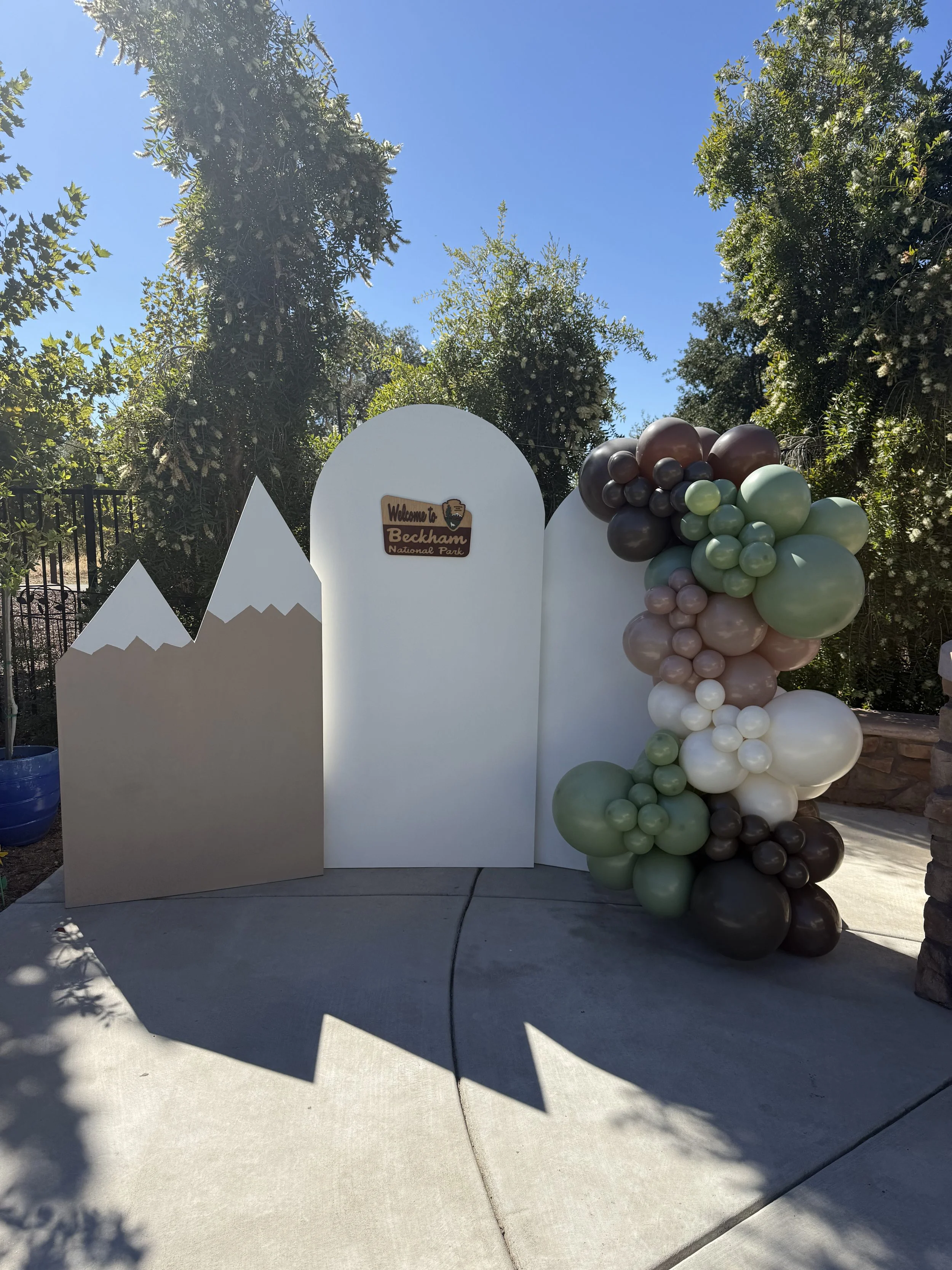 Decorative photo backdrop with mountain and hill elements, a sign that says "Welcome to Beckham National Park," and a cluster of green, white, pink, and brown balloons, set outdoors on a sunny day.
