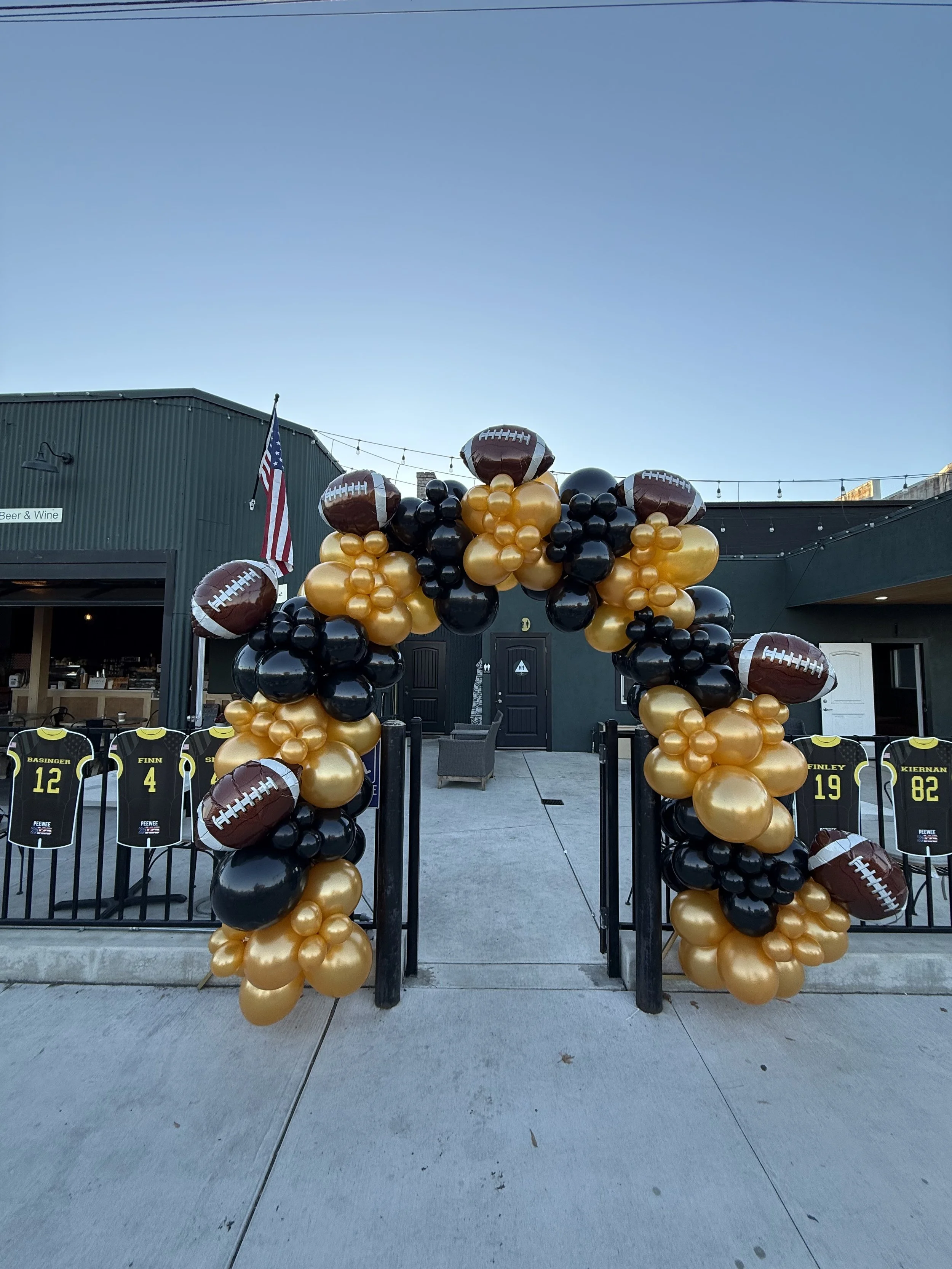 Football-themed balloon arch with black and gold balloons and football-shaped balloons over an outdoor patio with jerseys displayed and string lights.