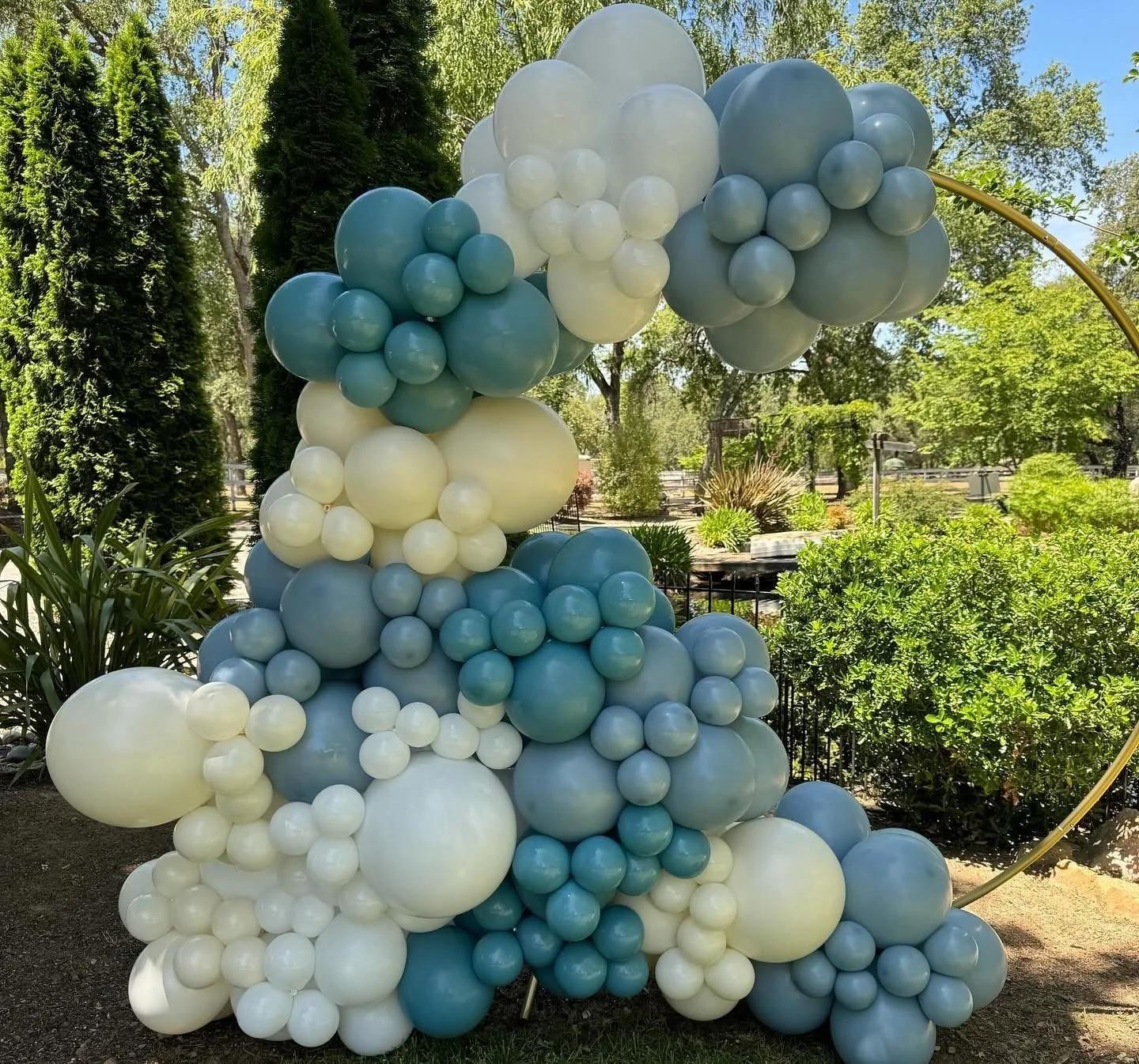 Arrangement of blue, white, and beige balloons in a cluster outdoors under trees.