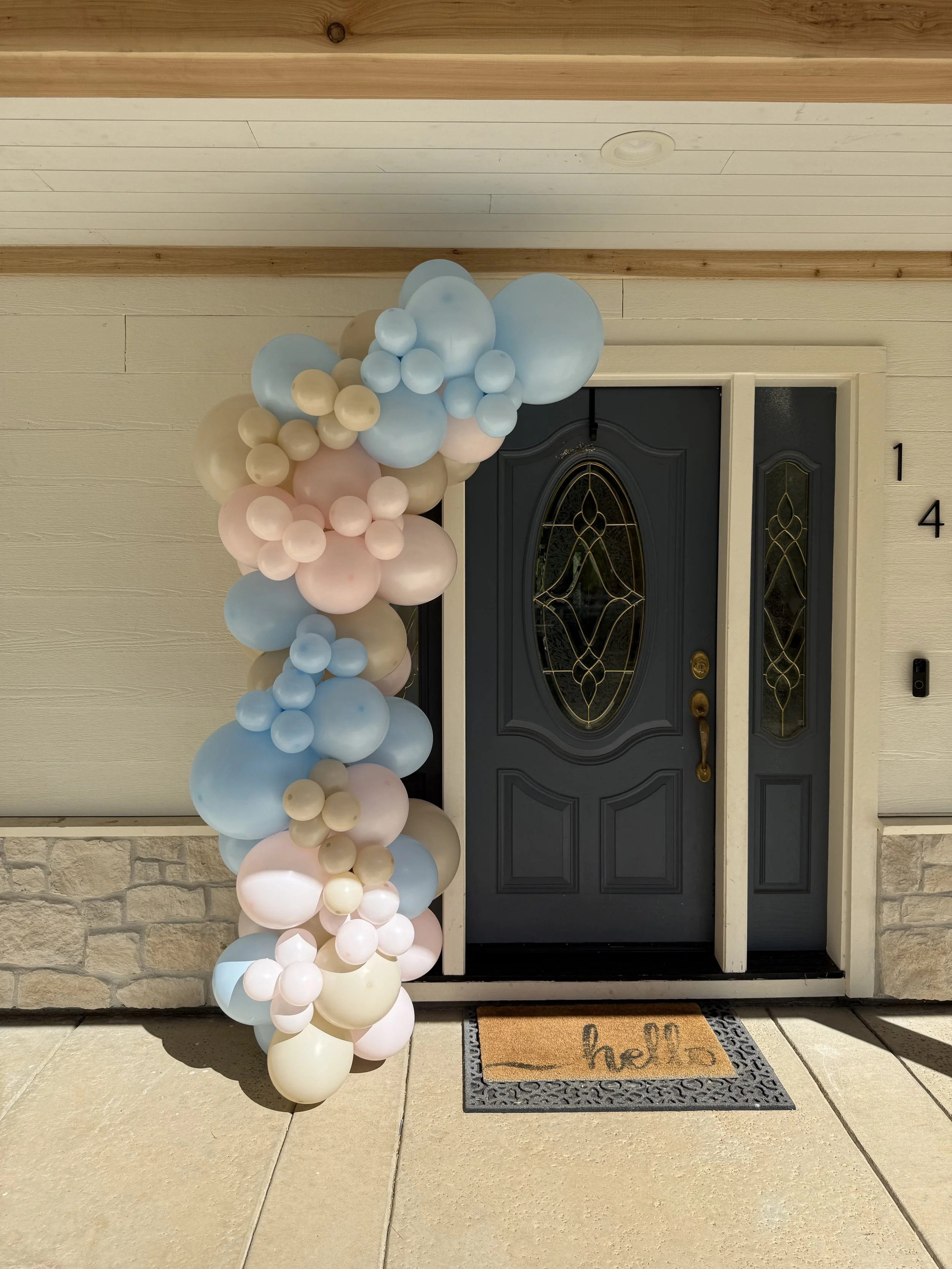 Balloon decoration in pastel colors placed beside a front door with a welcome mat that has the word 'hello' on it.