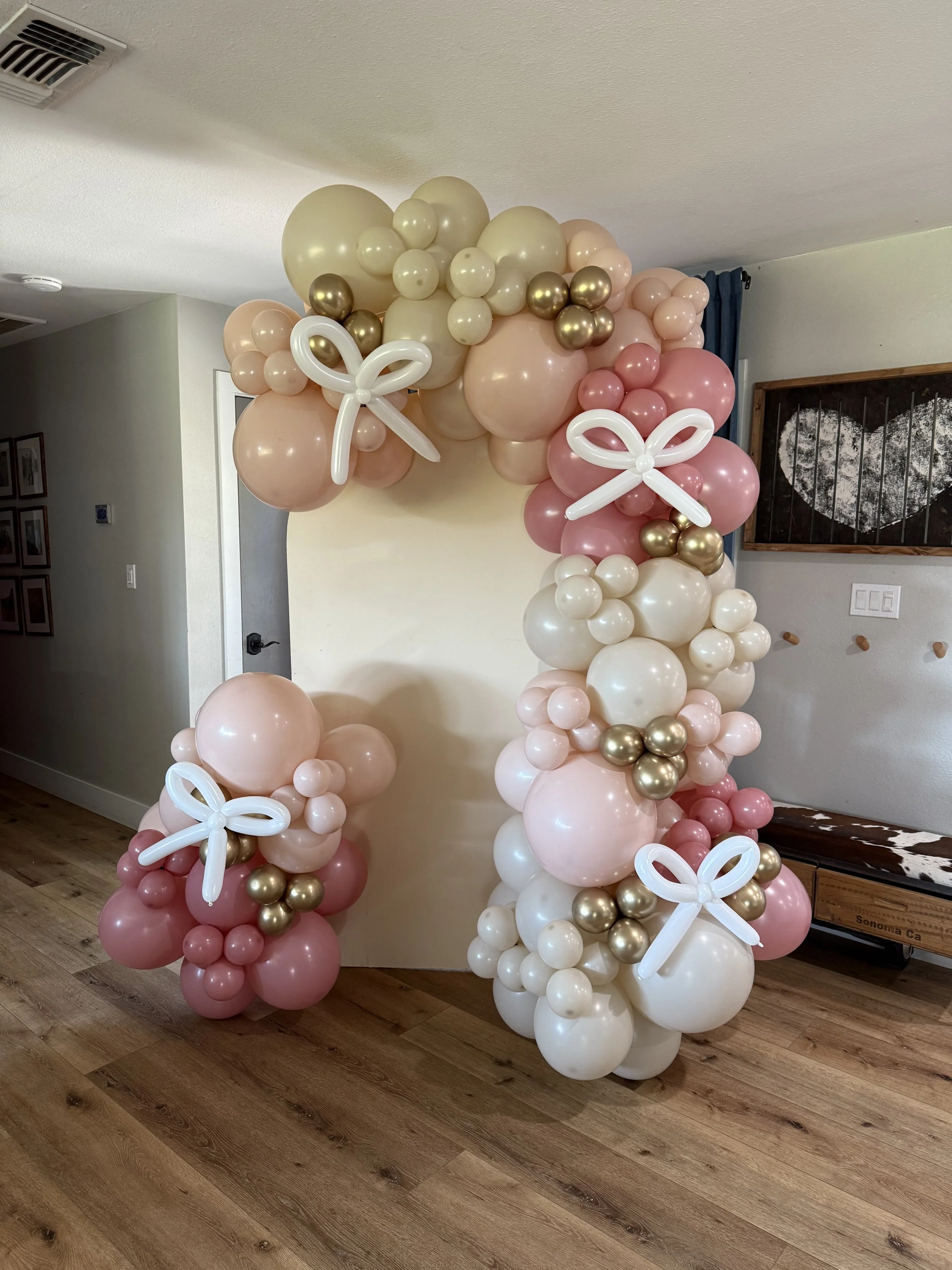 Decorative balloon arch with pink, white, gold, and cream-colored balloons and white balloon bows, set up indoors on a wooden floor.