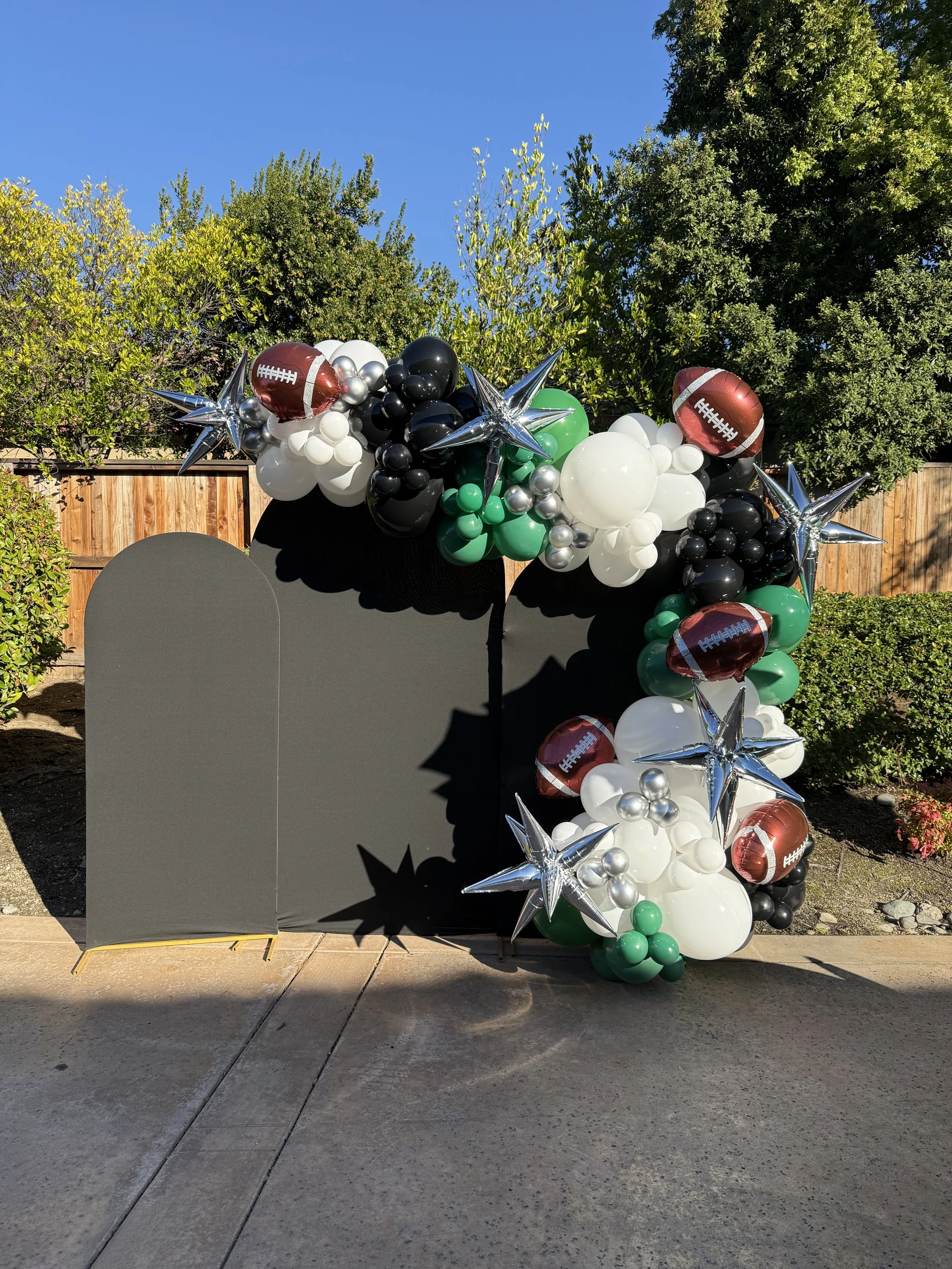 Football-themed balloon arrangement with black, white, green, silver, and red balloons, featuring football-shaped balloons and star-shaped metallic balloons, set against a black backdrop outdoors.