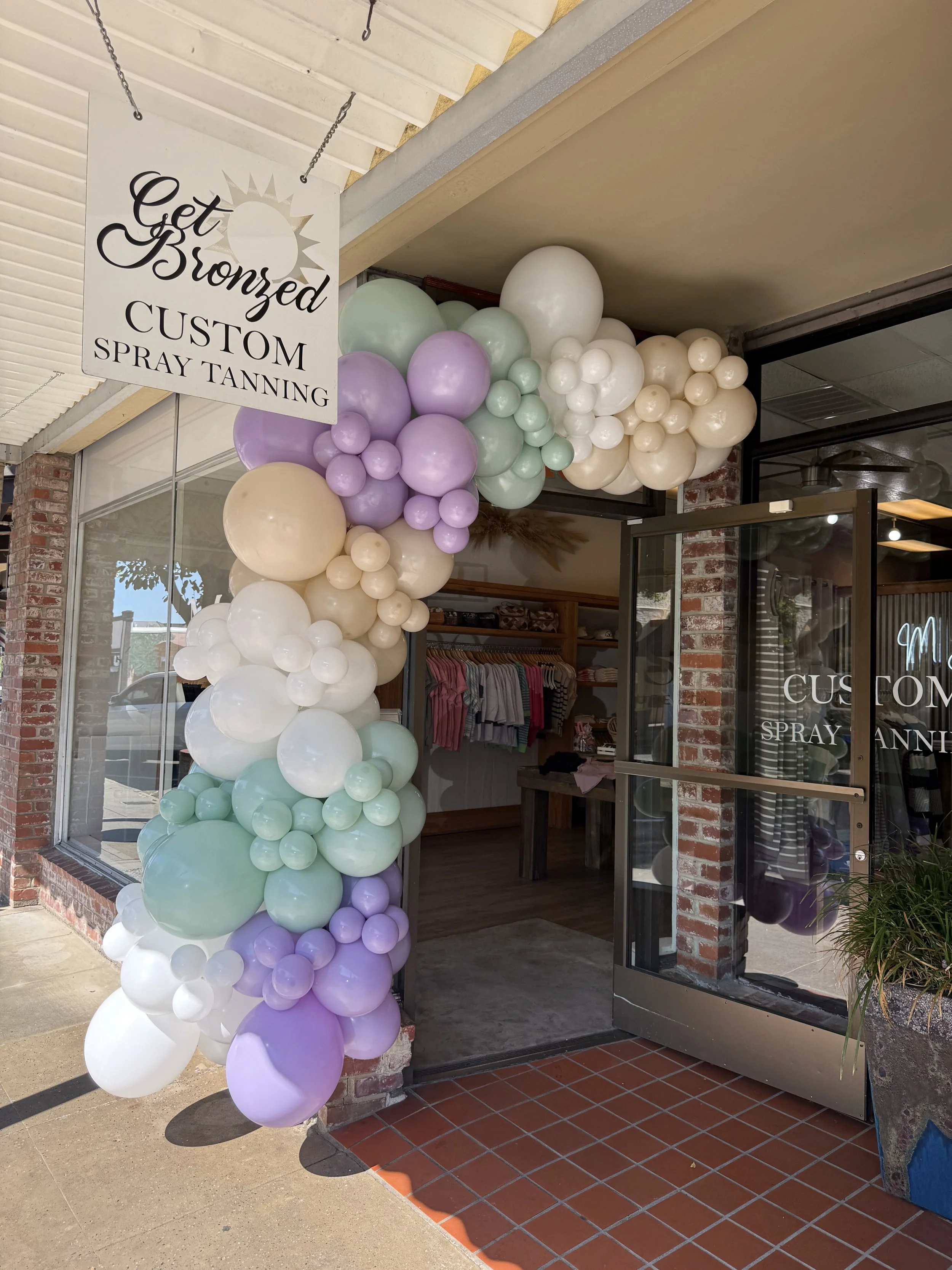 Entrance of a storefront decorated with pastel-colored balloons in purple, mint, white, and cream, with a sign hanging that reads 'Get Bronzed Custom Spray Tanning'.