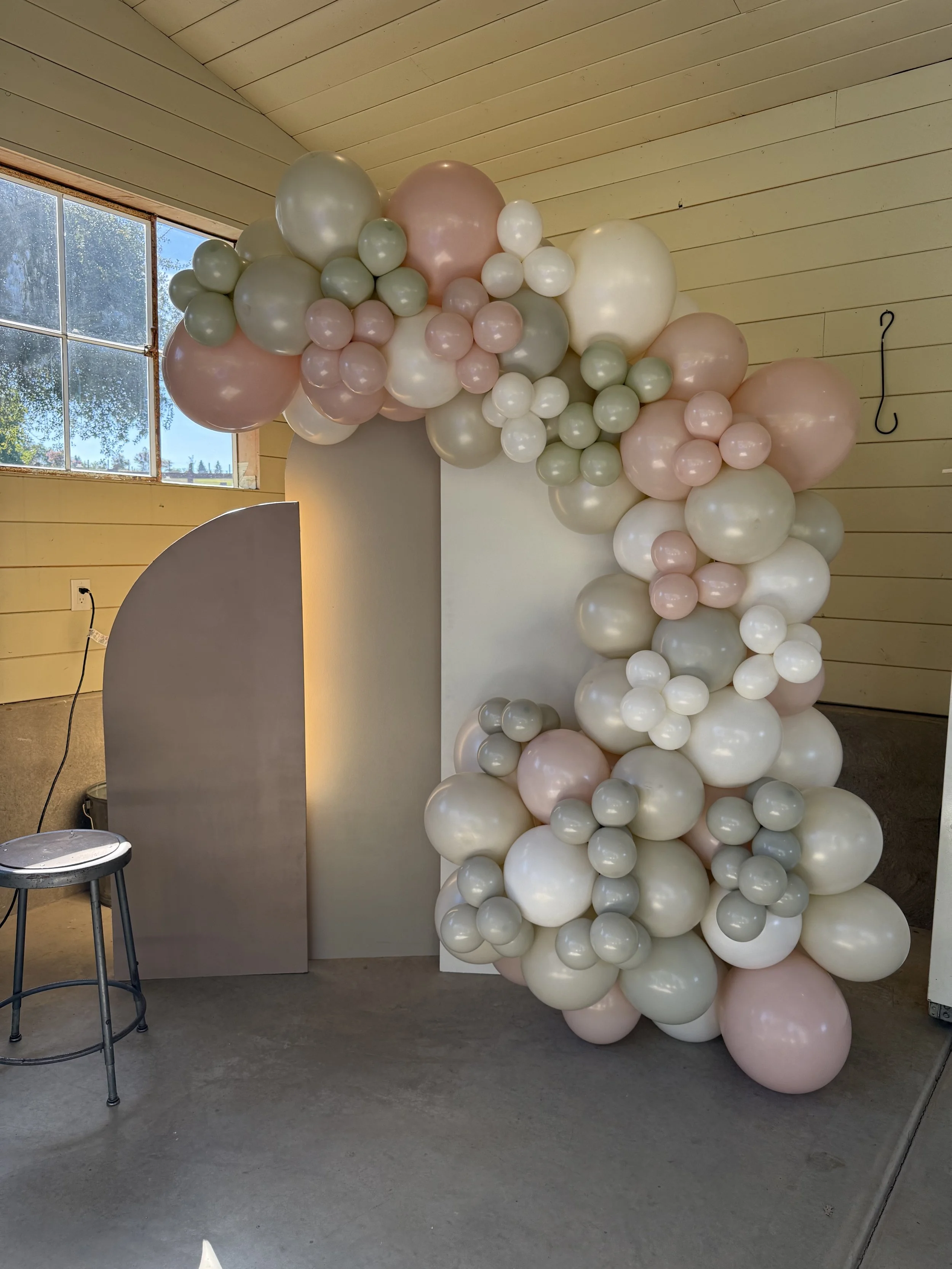 Balloon arch with pink, white, and grey balloons in a room with yellow wooden walls and a large window.