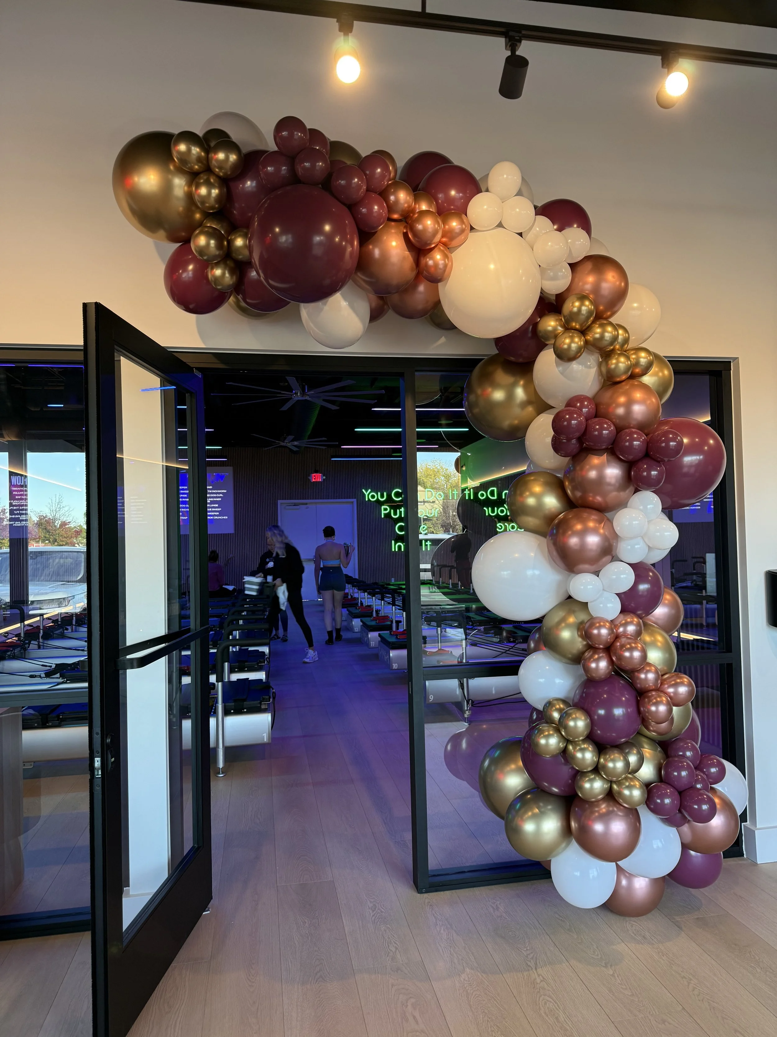 Colorful balloon arch with gold, white, and maroon balloons decorating the entrance of a fitness or dance studio.