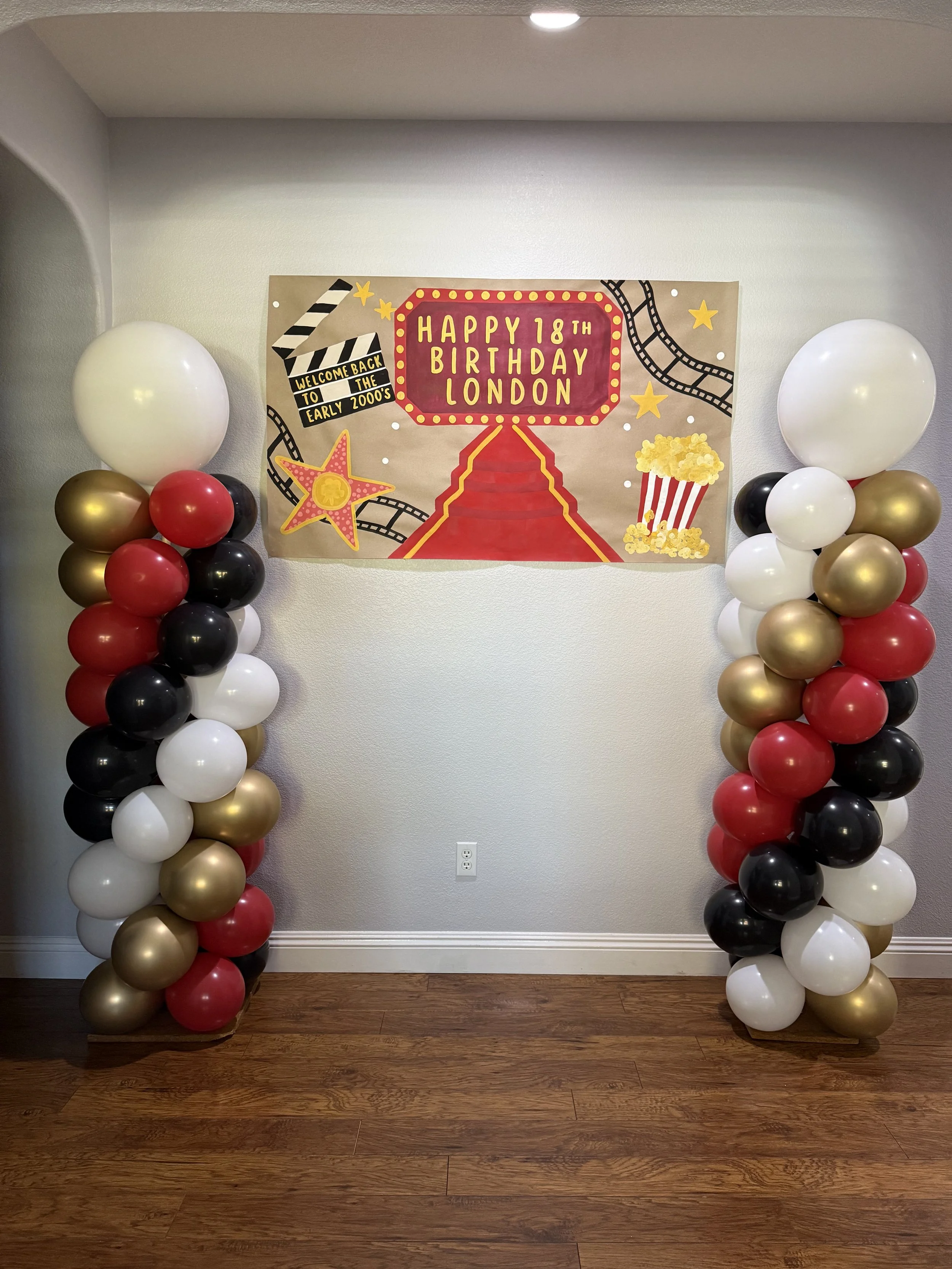 Decorative birthday celebration setup with a large sign that says 'Happy 18th Birthday London' with an illustration of a red carpet, popcorn, and stars, flanked by two columns of balloons in white, black, red, and gold.