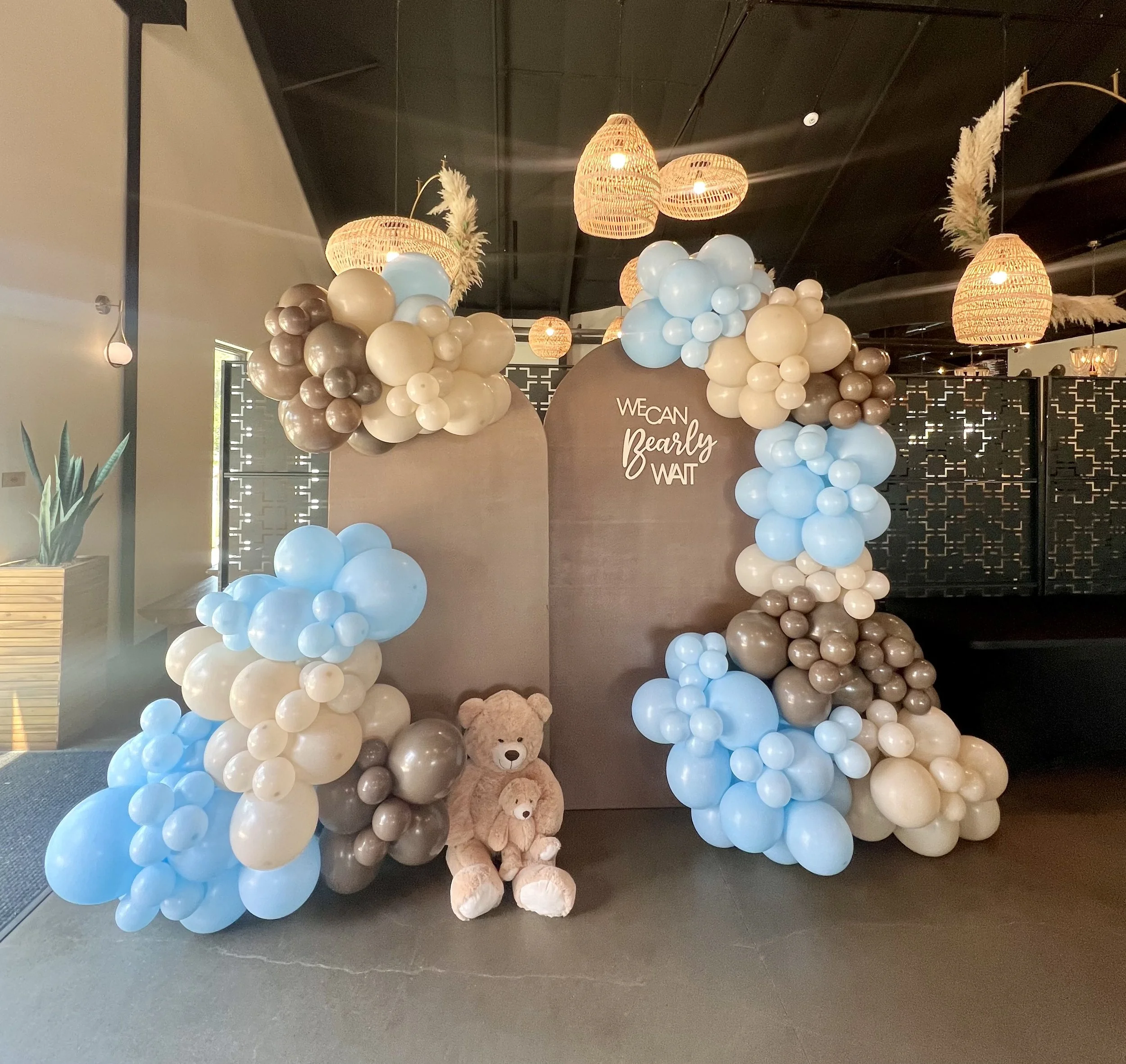 Decorative balloon arch with blue, beige, and gray balloons, teddy bears, and a sign that reads 'We can, Bearly-wait' in an indoor setting.