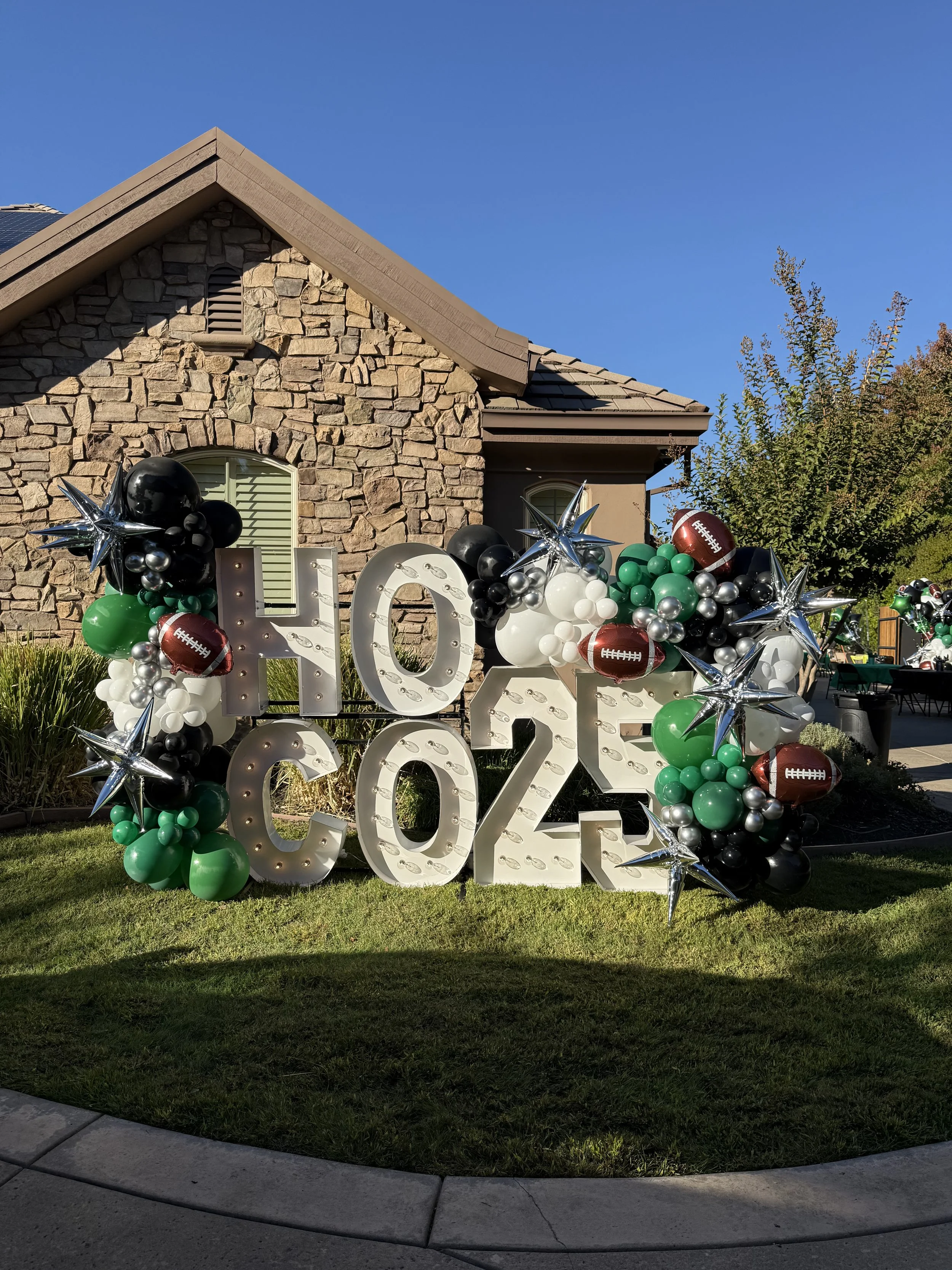large sign with the numbers 2023, decorated with balloons in black, white, green, and silver, and football-shaped balloons, placed outside a house with a stone facade and a lawn, under a clear blue sky.