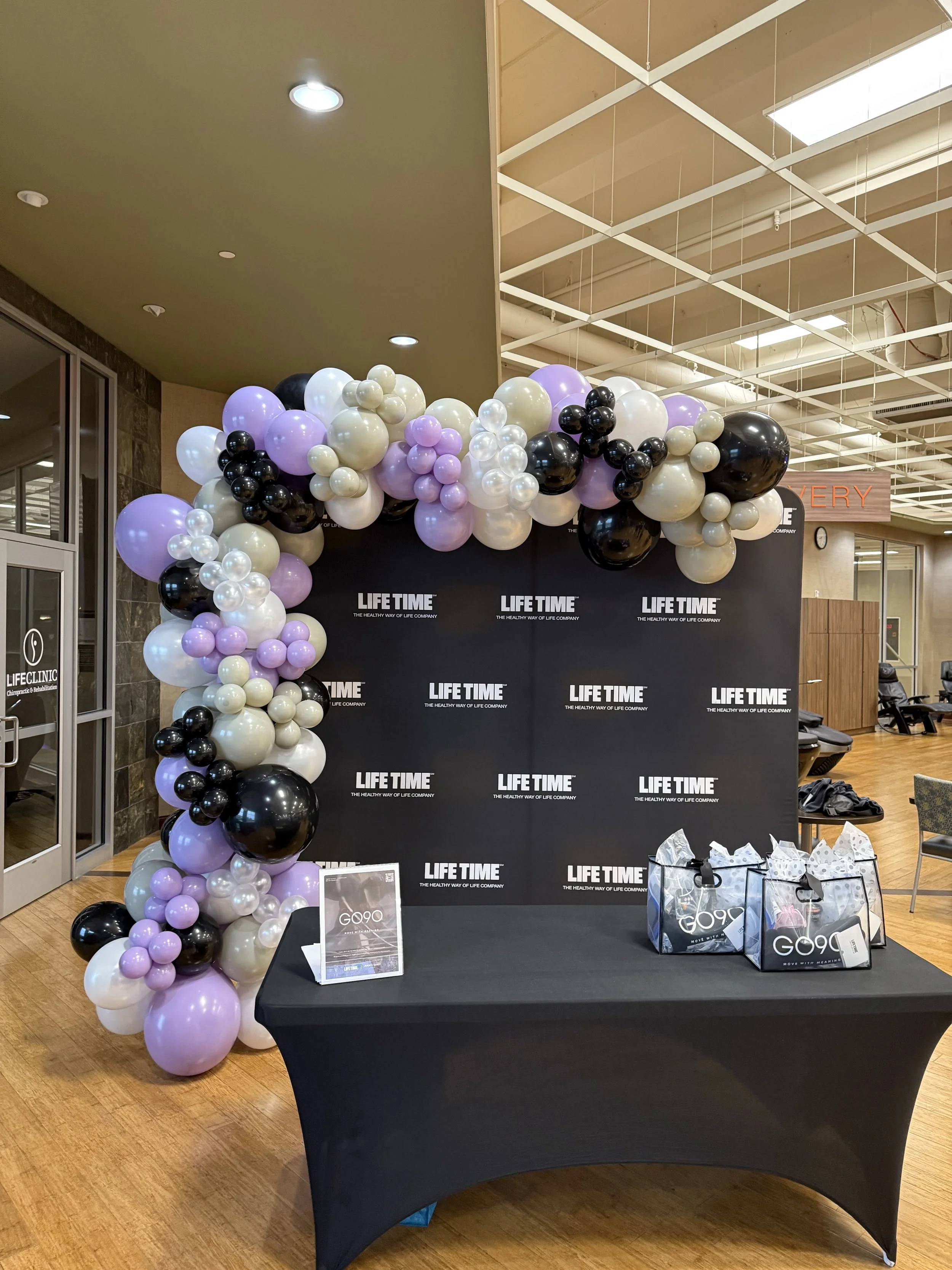 Decorative balloon arch with purple, black, white, and pearl-colored balloons at a healthcare event, in front of a black backdrop with 'LIFE TIME' branding, and a table with gift bags.