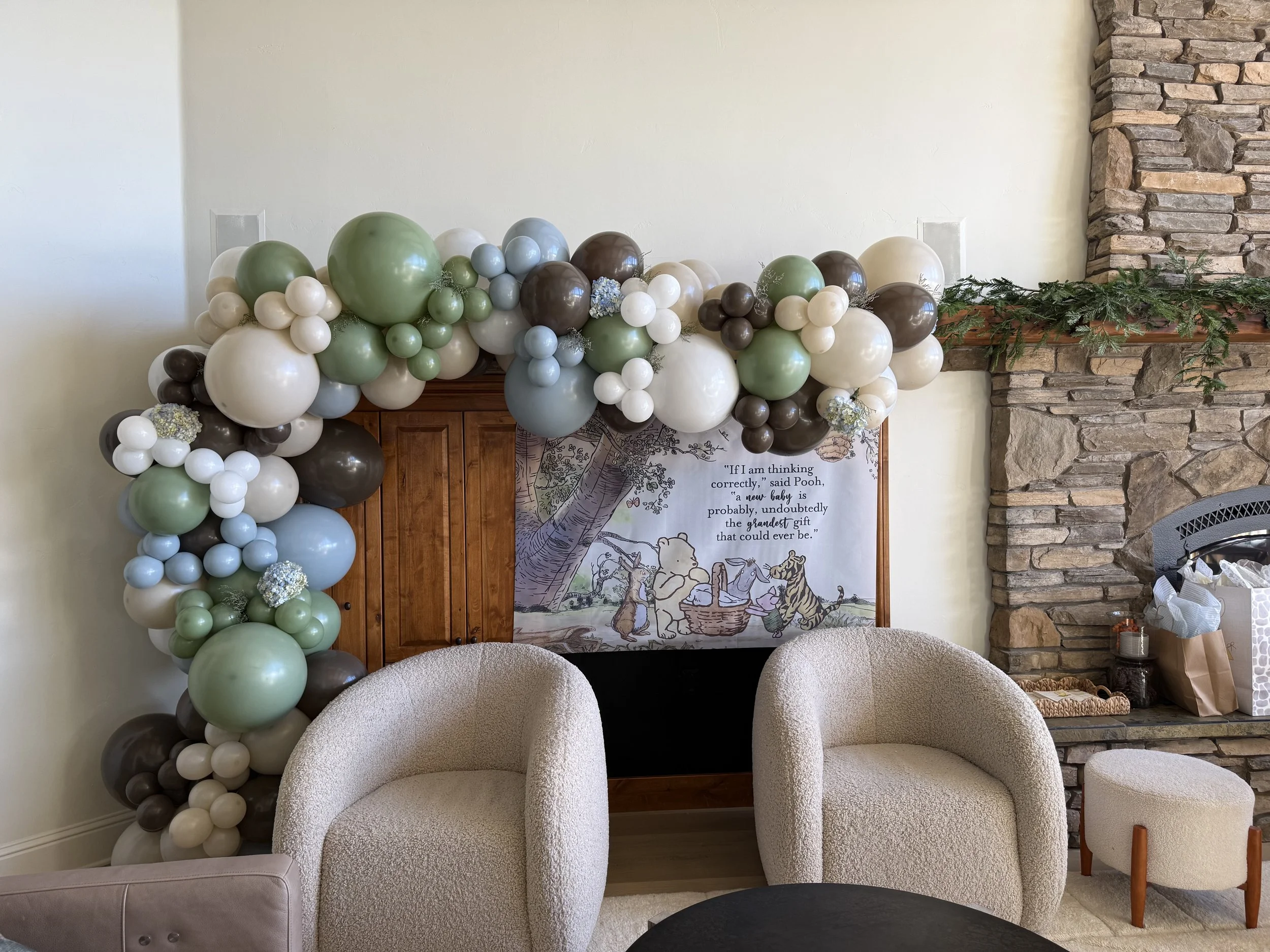 Balloon arch with shades of white, green, blue, gray, and brown balloons over a fireplace and wooden cabinet in a living room.
